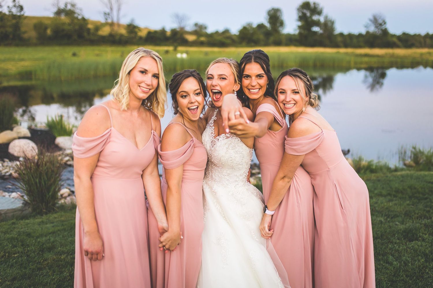 Bridesmaids in blush pink dresses pose with bride by scenic lake backdrop.