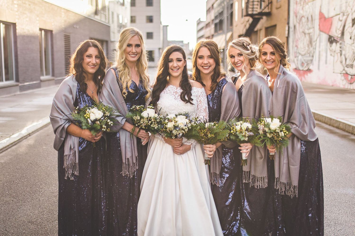 Bridesmaids in matching outfits gather around bride on city sidewalk.