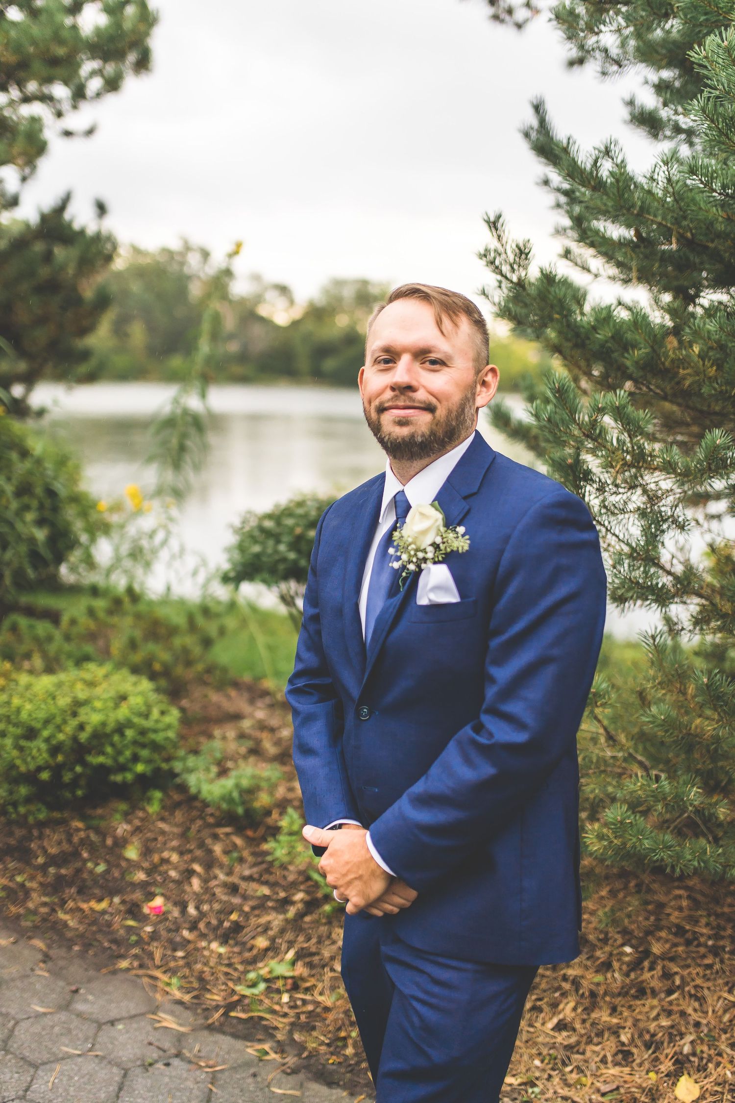 Groom in navy suit poses by lakeside garden wearing boutonniere and classic tie.