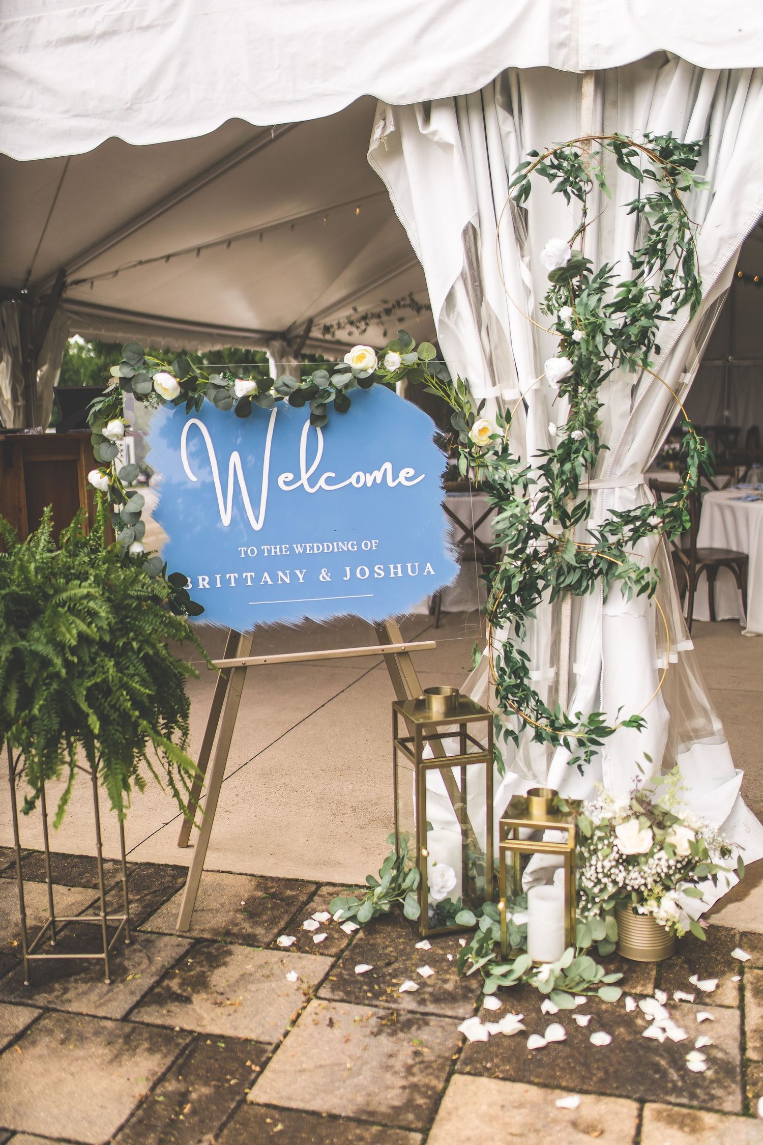 Wedding tent entrance decorated with white drapes, greenery and a blue welcome sign.
