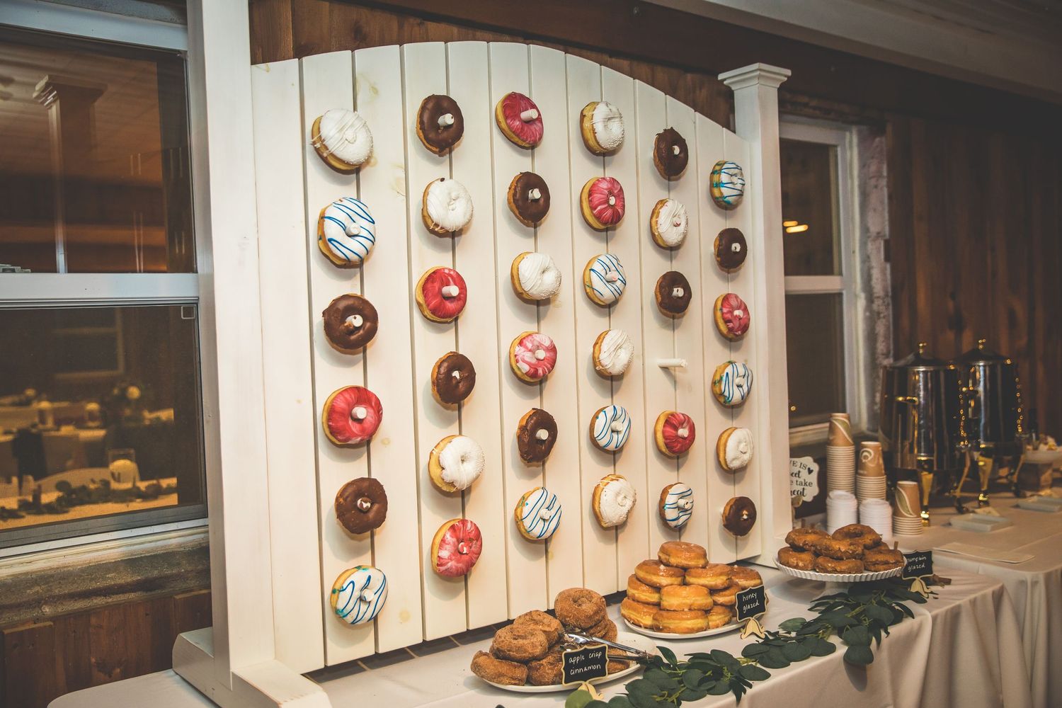 Creative donut wall display at rustic wedding reception features colorful pastries on wooden board.