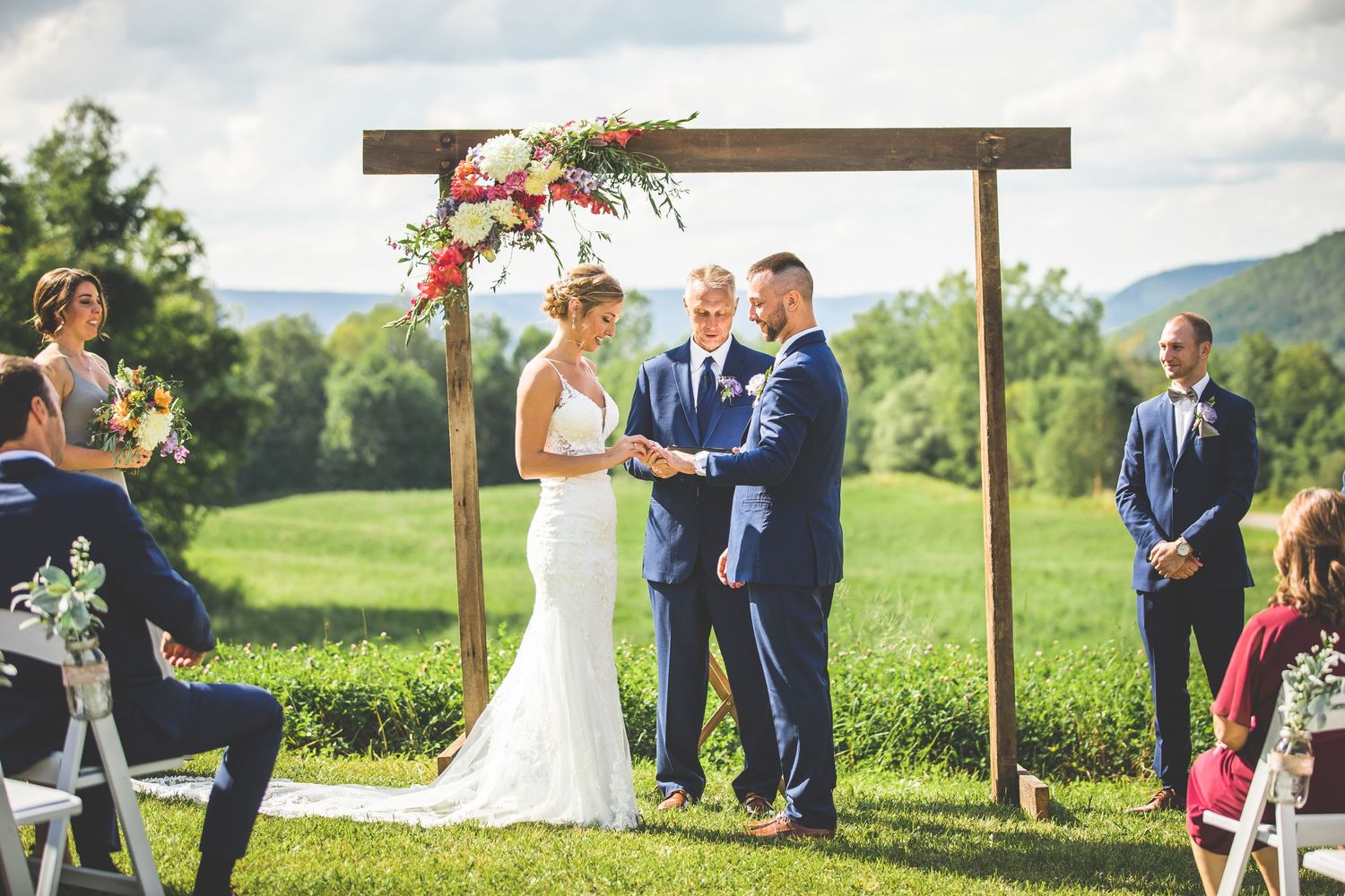 Wedding ceremony taking place under wooden pergola with mountain views and white chairs for guests.