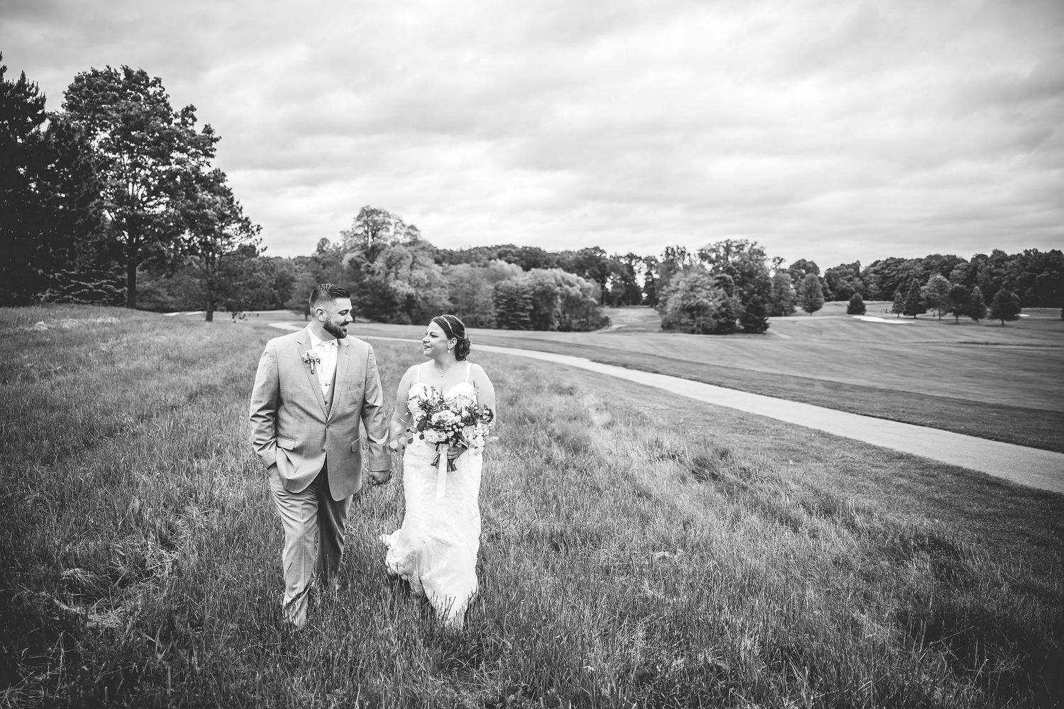 A black and white panoramic view of a couple walking through a scenic meadow on their wedding day.