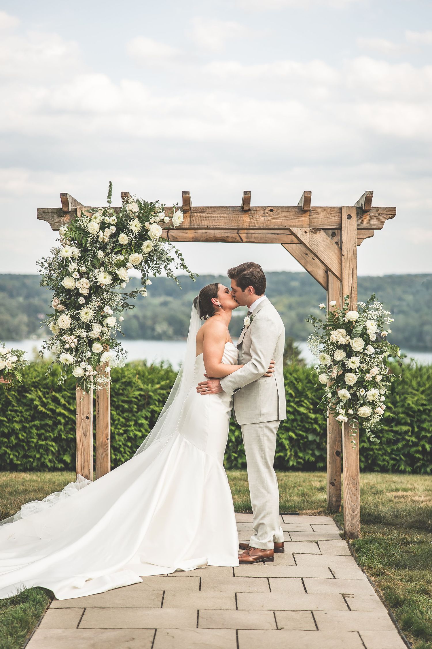 Newlyweds share intimate moment under floral-decorated wooden pergola with water view.