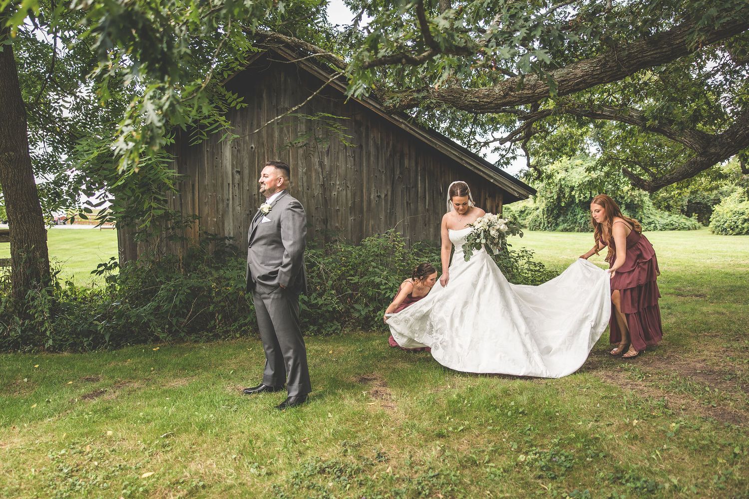 Wedding party gathers near old wooden barn for outdoor photo session.