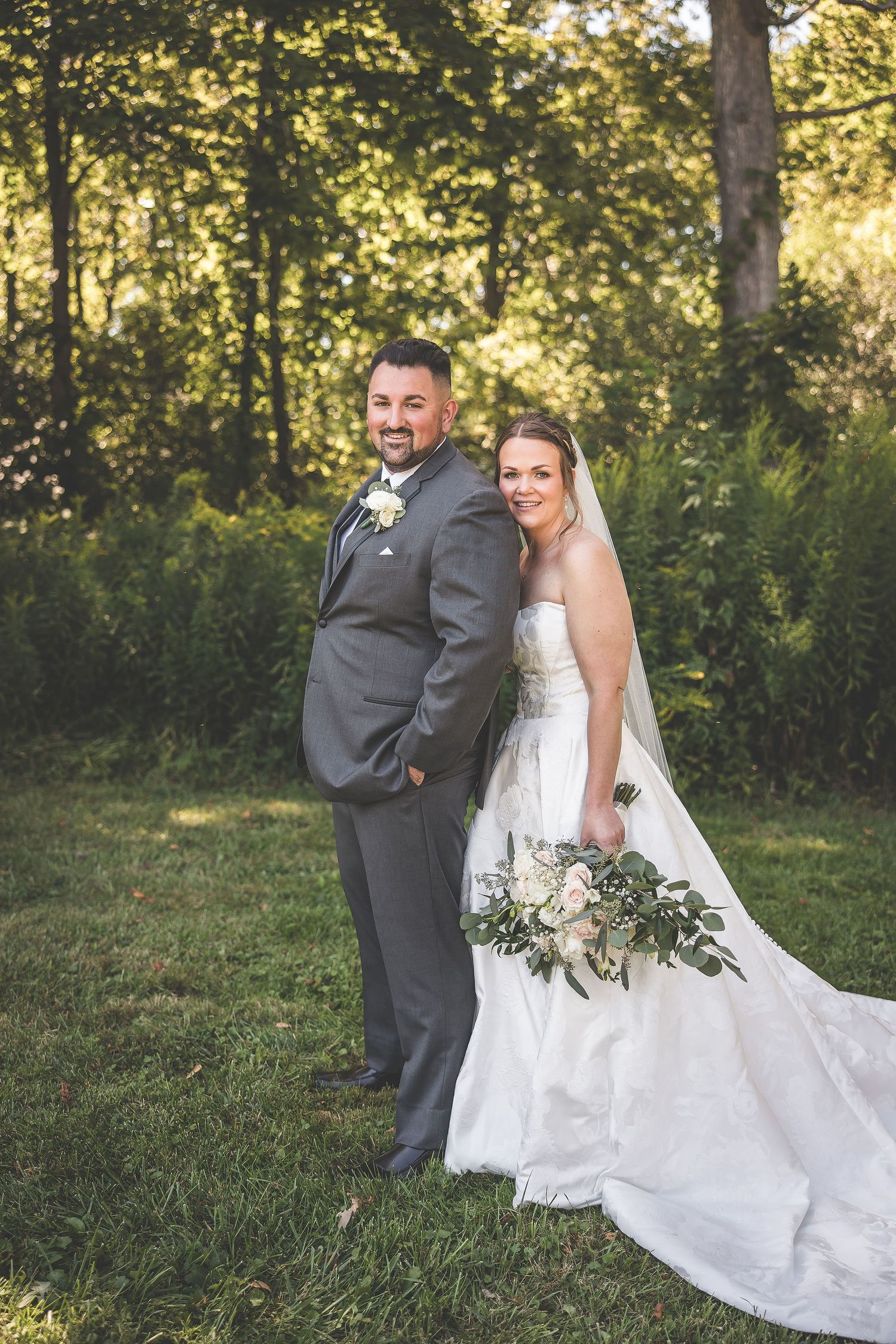 Wedding couple poses in gray suit and white gown with flowing train in sunlit garden setting.