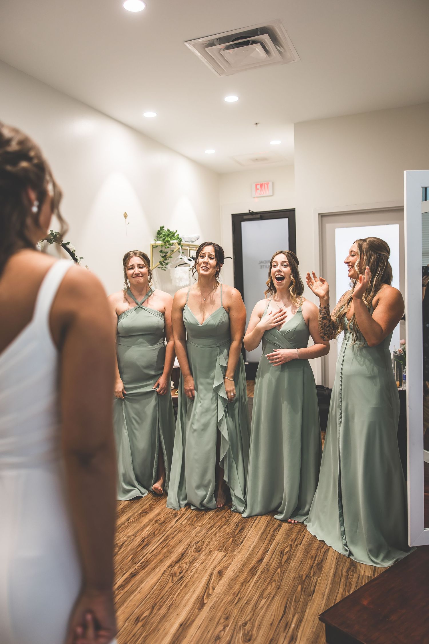 Bridesmaids in matching sage green dresses share excited reactions in a bright indoor space.