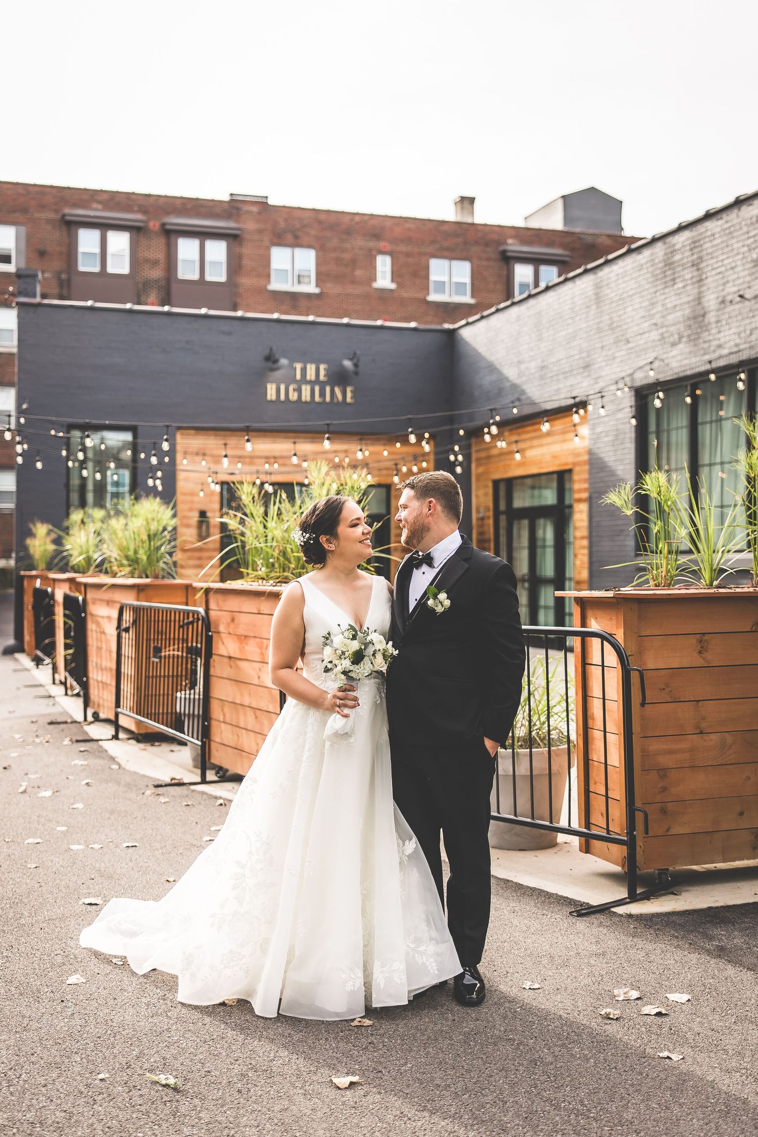 Wedding couple poses against modern brick and wood backdrop with string lights at urban venue.