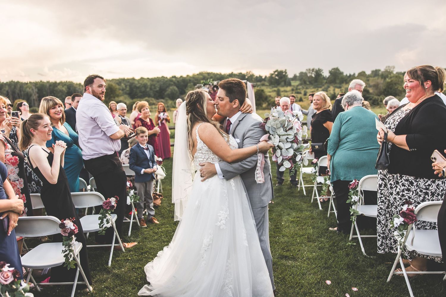 Wedding celebration on a scenic hillside with guests seated in white chairs during an outdoor ceremony.