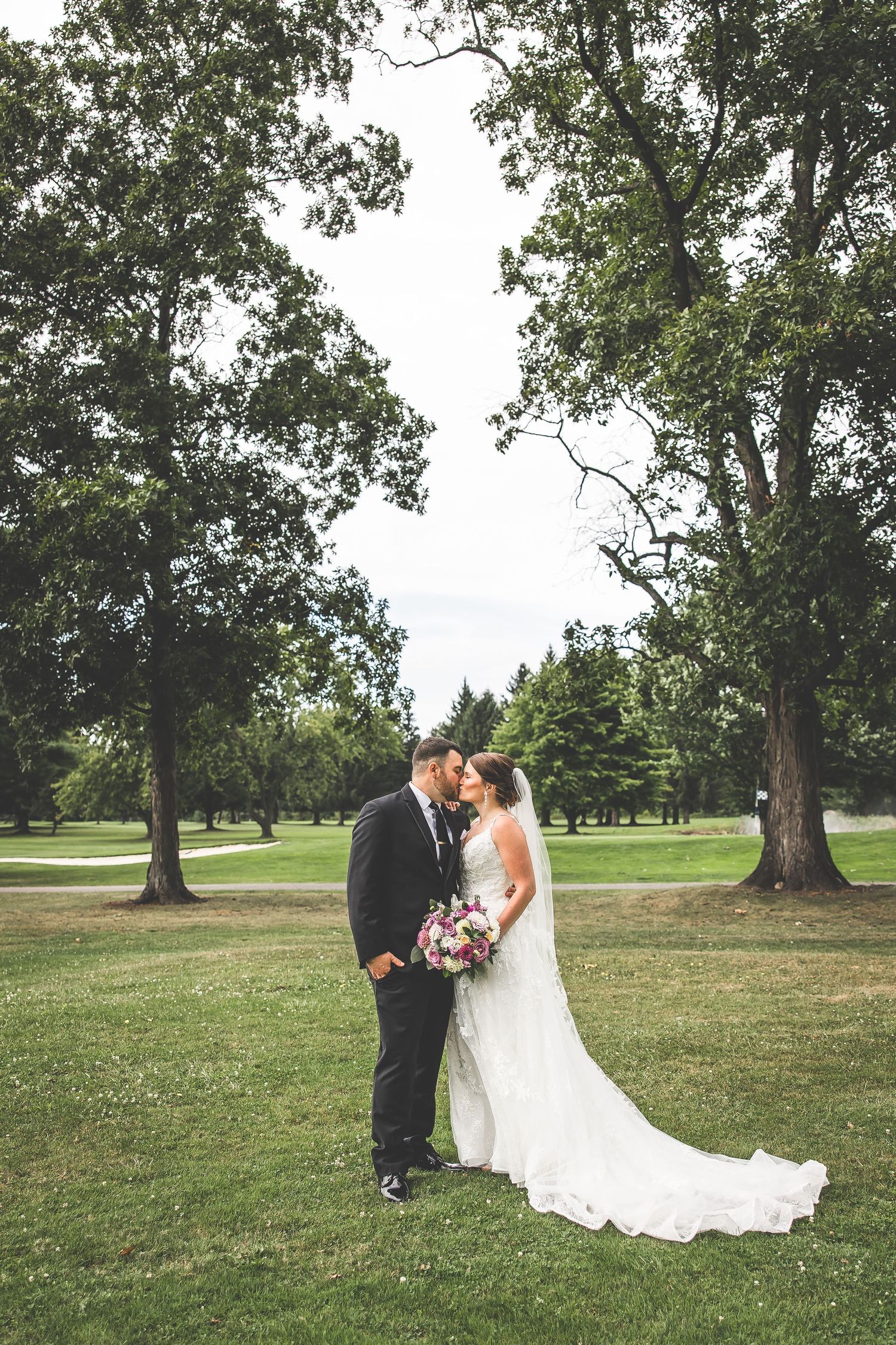 Wedding couple poses on a sprawling lawn with majestic oak trees creating a natural outdoor backdrop.