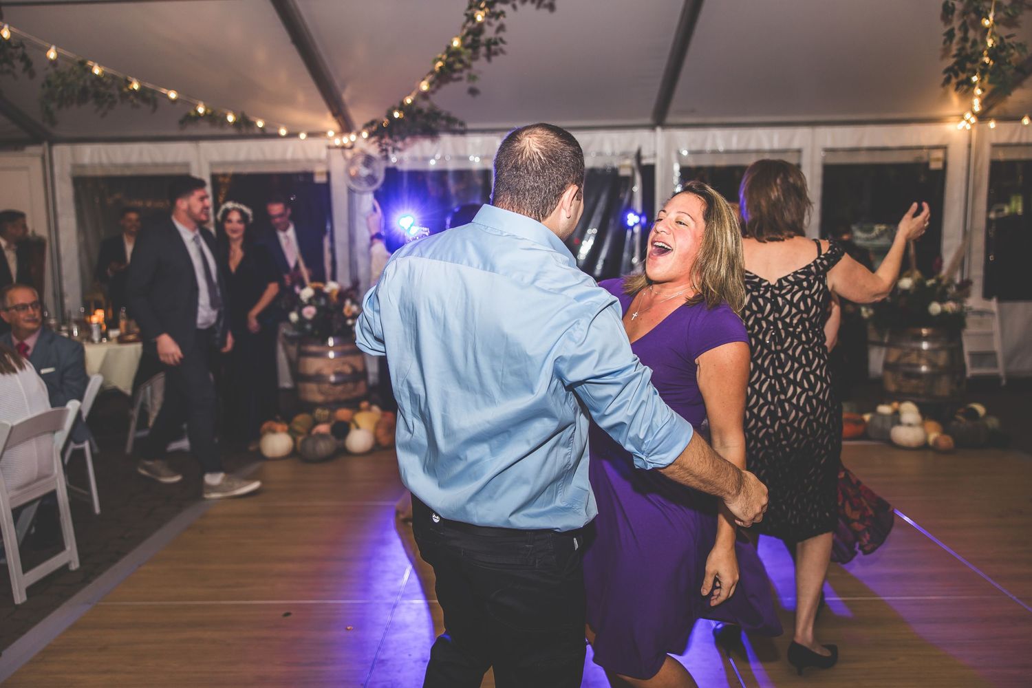 Wedding guests enjoy dancing at a reception with purple lighting and white tent decor.