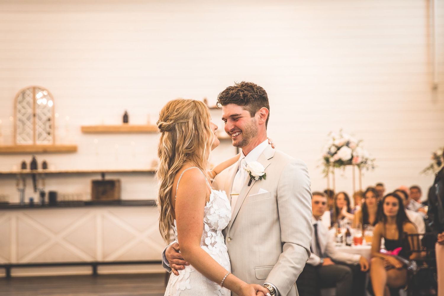 Newlyweds share a romantic dance in a light-filled reception hall with guests looking on.