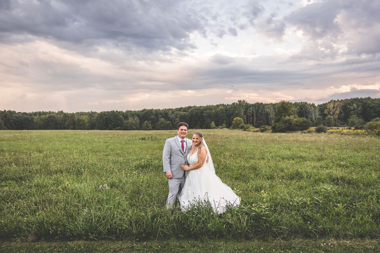 Newlyweds pose together in open field during sunset with cloudy sky backdrop.