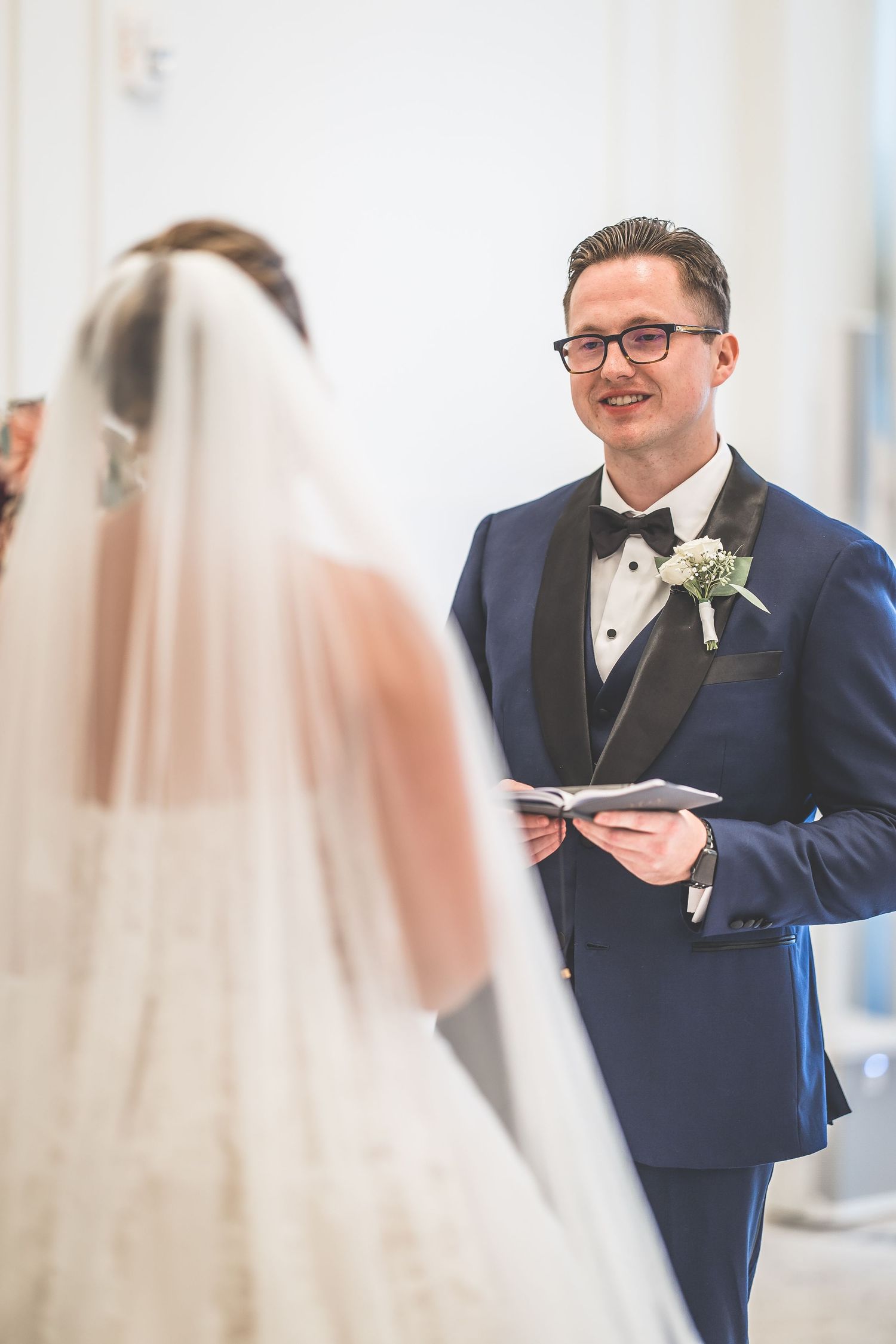 A couple exchanging vows during their wedding ceremony with the groom in a navy tuxedo reading from a booklet.
