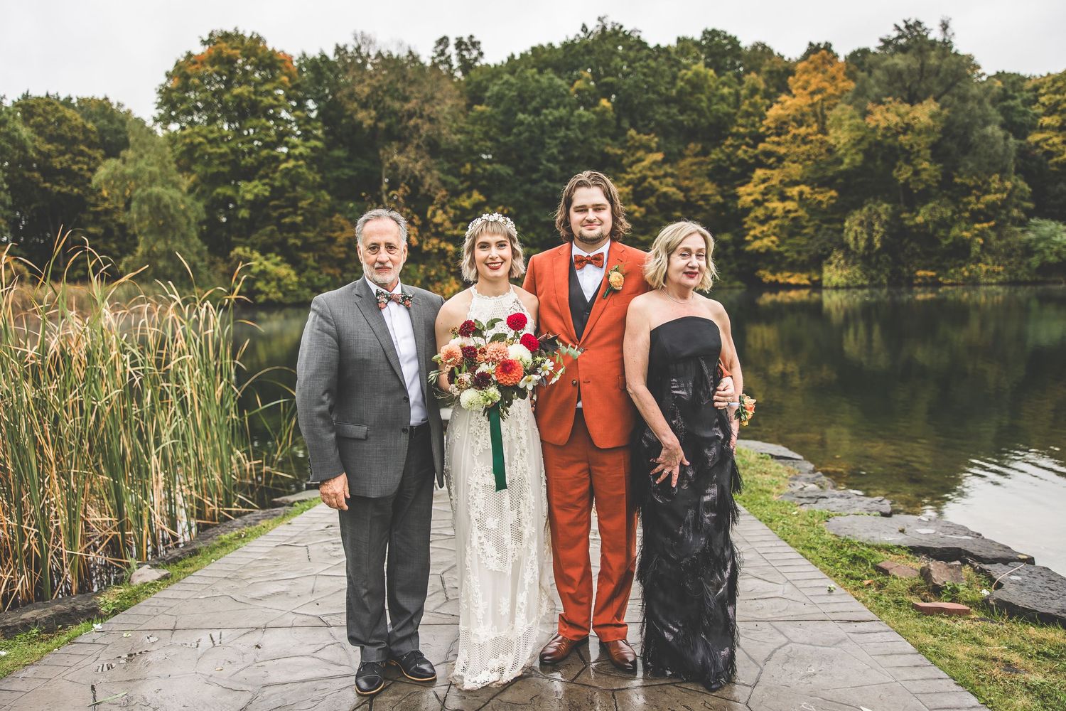 Small wedding party poses together on stone path by tranquil lake with fall foliage.