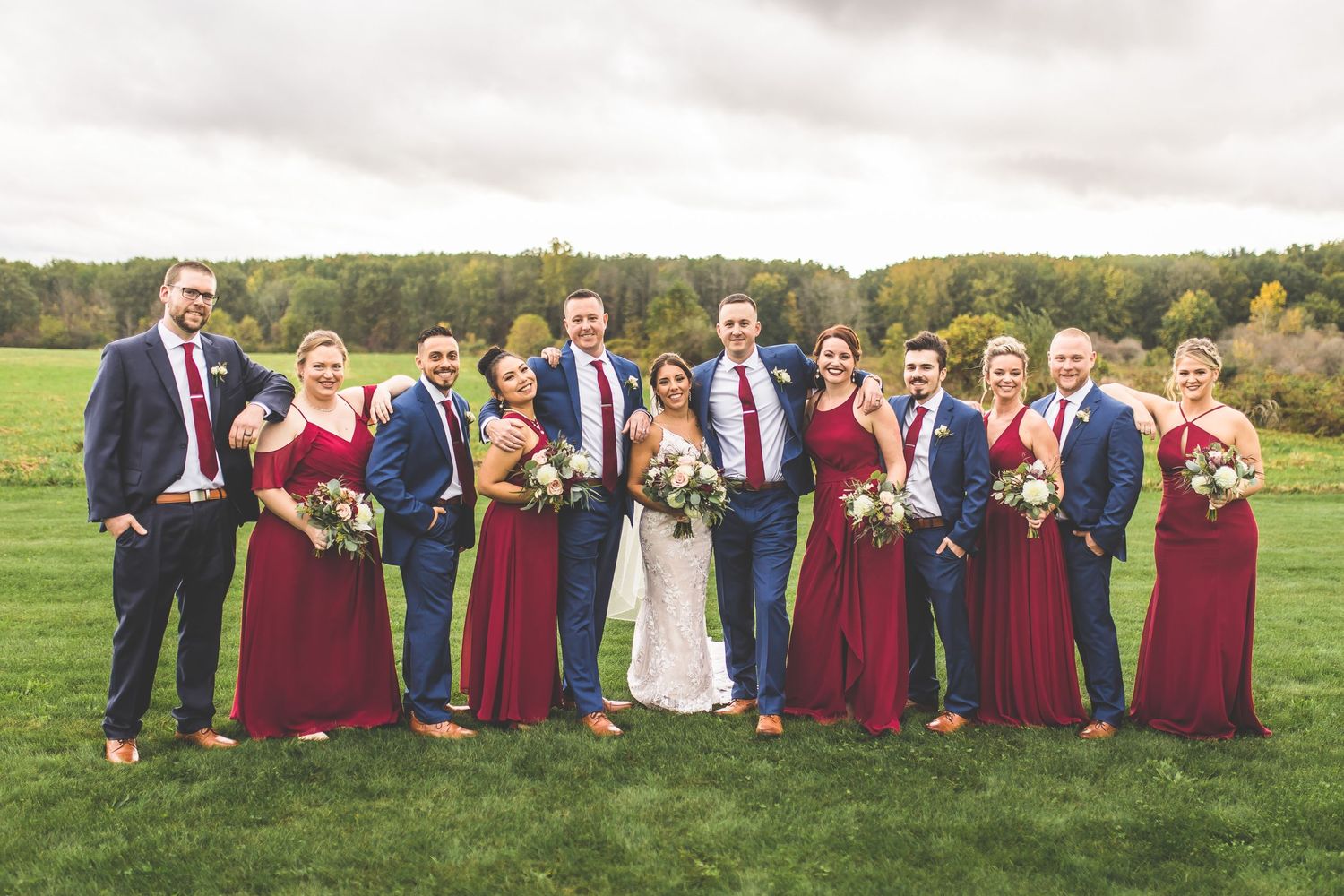 Wedding party lined up on grassy field with burgundy and navy color scheme against autumn backdrop.