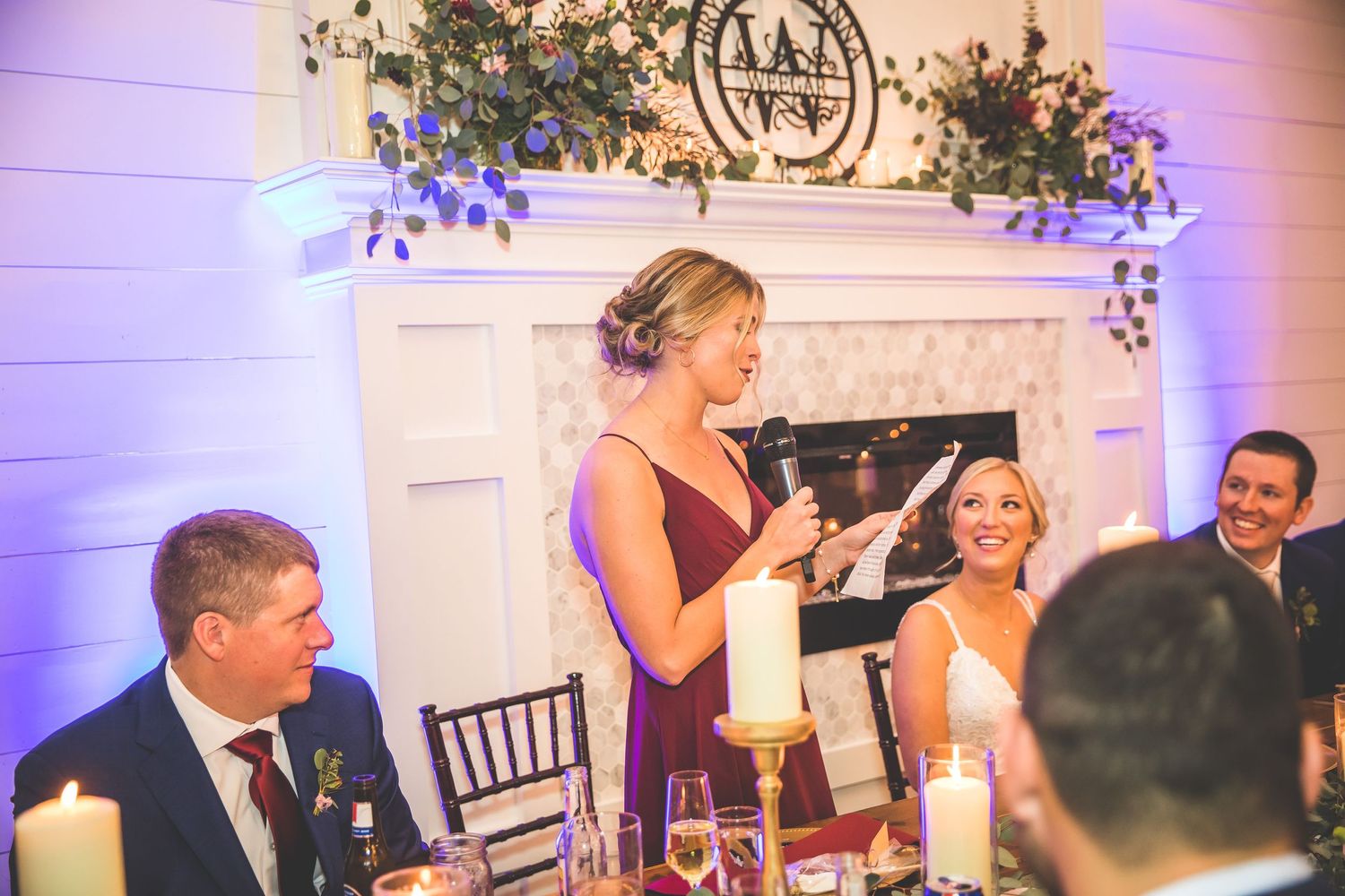 Wedding reception speech being given at an elegant venue with decorative fireplace and floral arrangements.