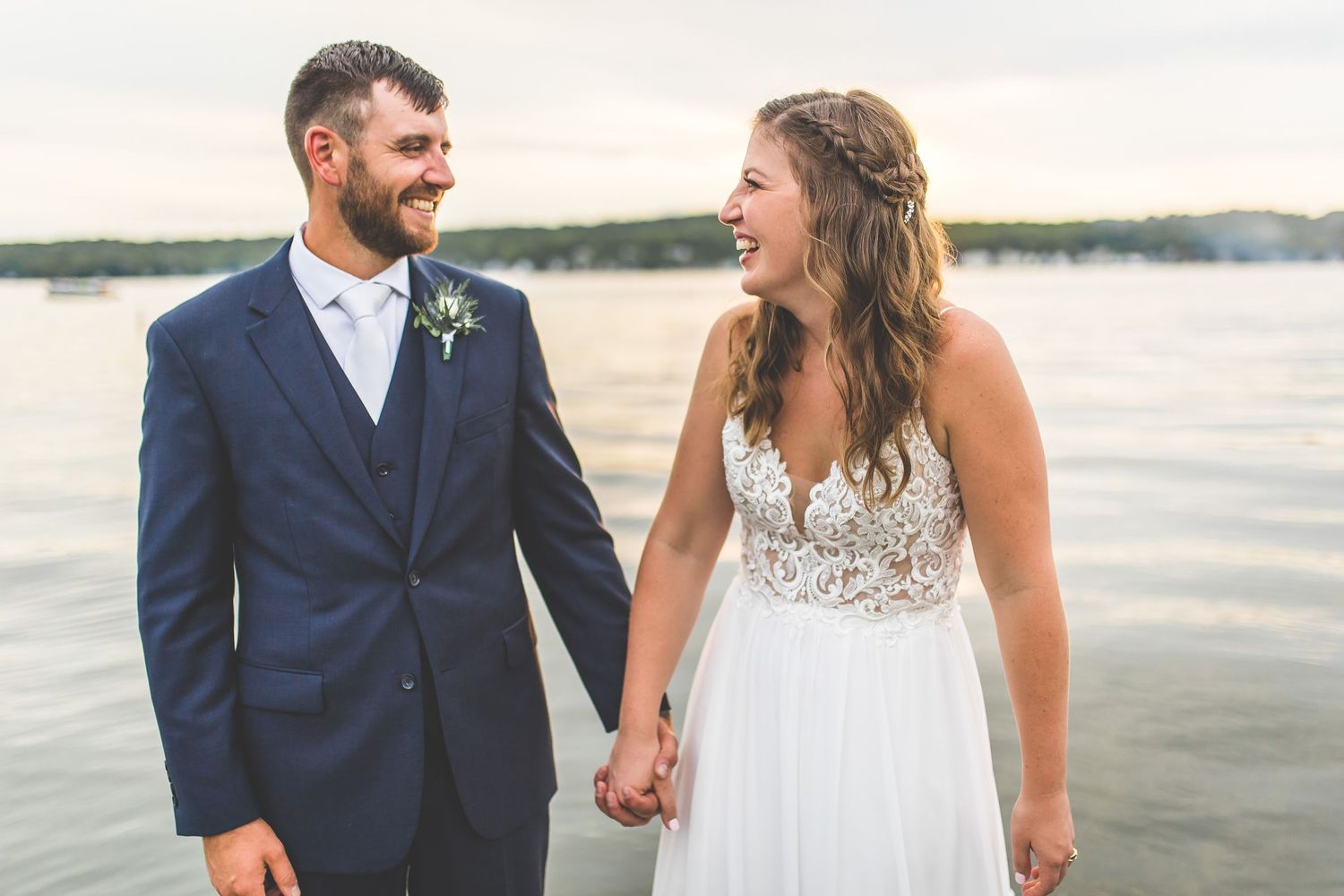 A couple shares a romantic moment at sunset on a lakeside dock wearing formal wedding attire.