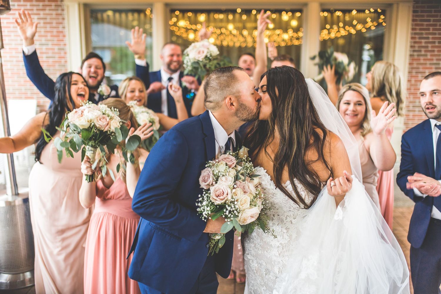 Wedding party celebrates with joyful cheers as newlyweds share a kiss under twinkling lights.