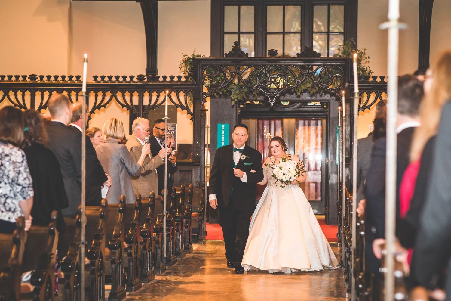 Wedding ceremony procession in a grand church aisle with wooden pews and decorative lighting.