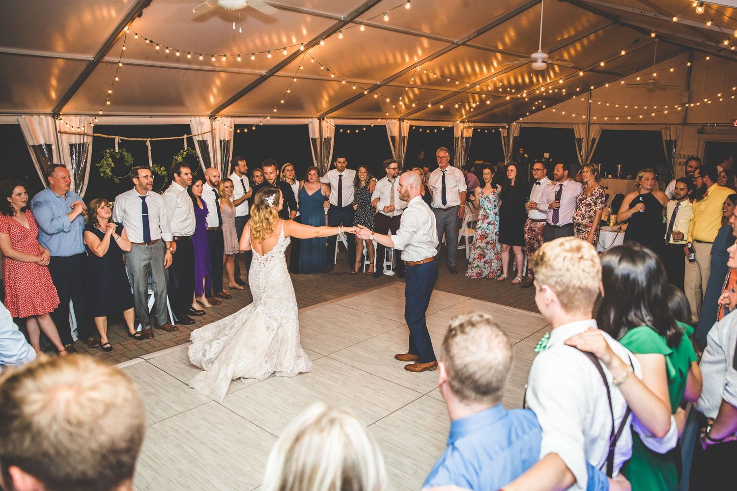 Wedding reception dance floor filled with guests under warm string lights and wooden beams.