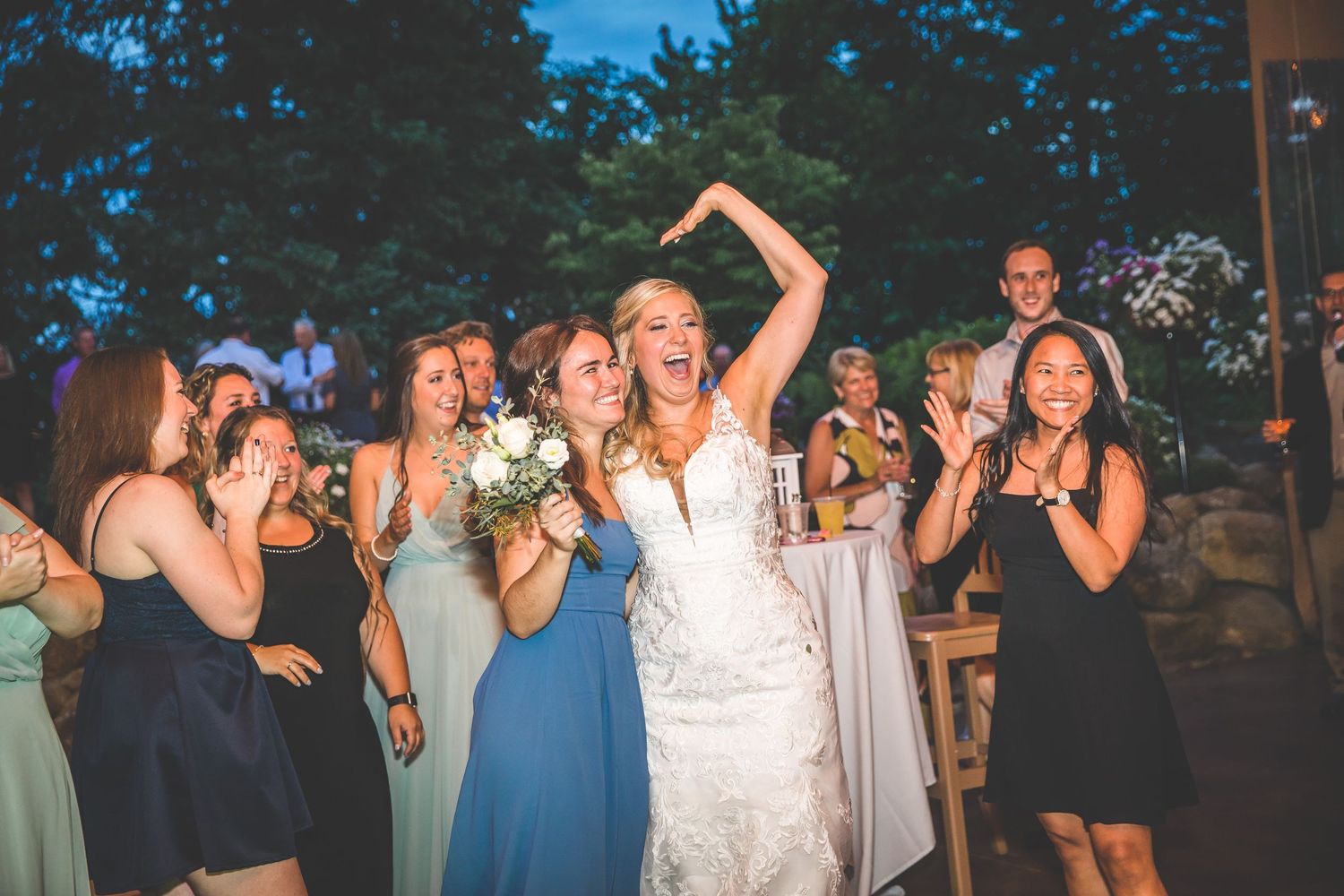 Wedding party celebrates outdoors at dusk with bridesmaids in various colored dresses surrounding the bride.