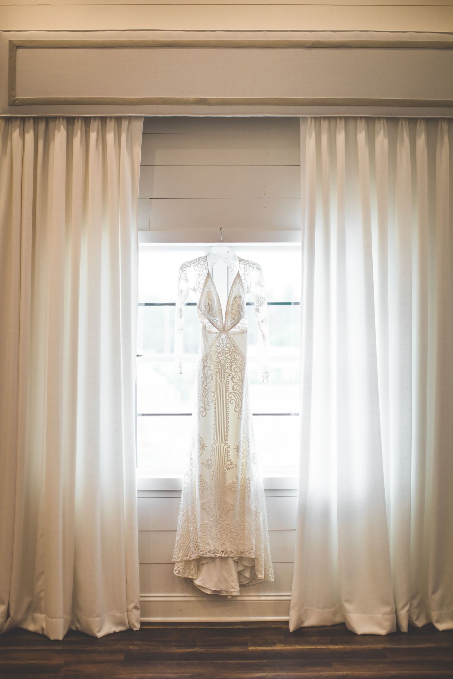 Elegant lace wedding dress hanging in window between white curtains in natural light.