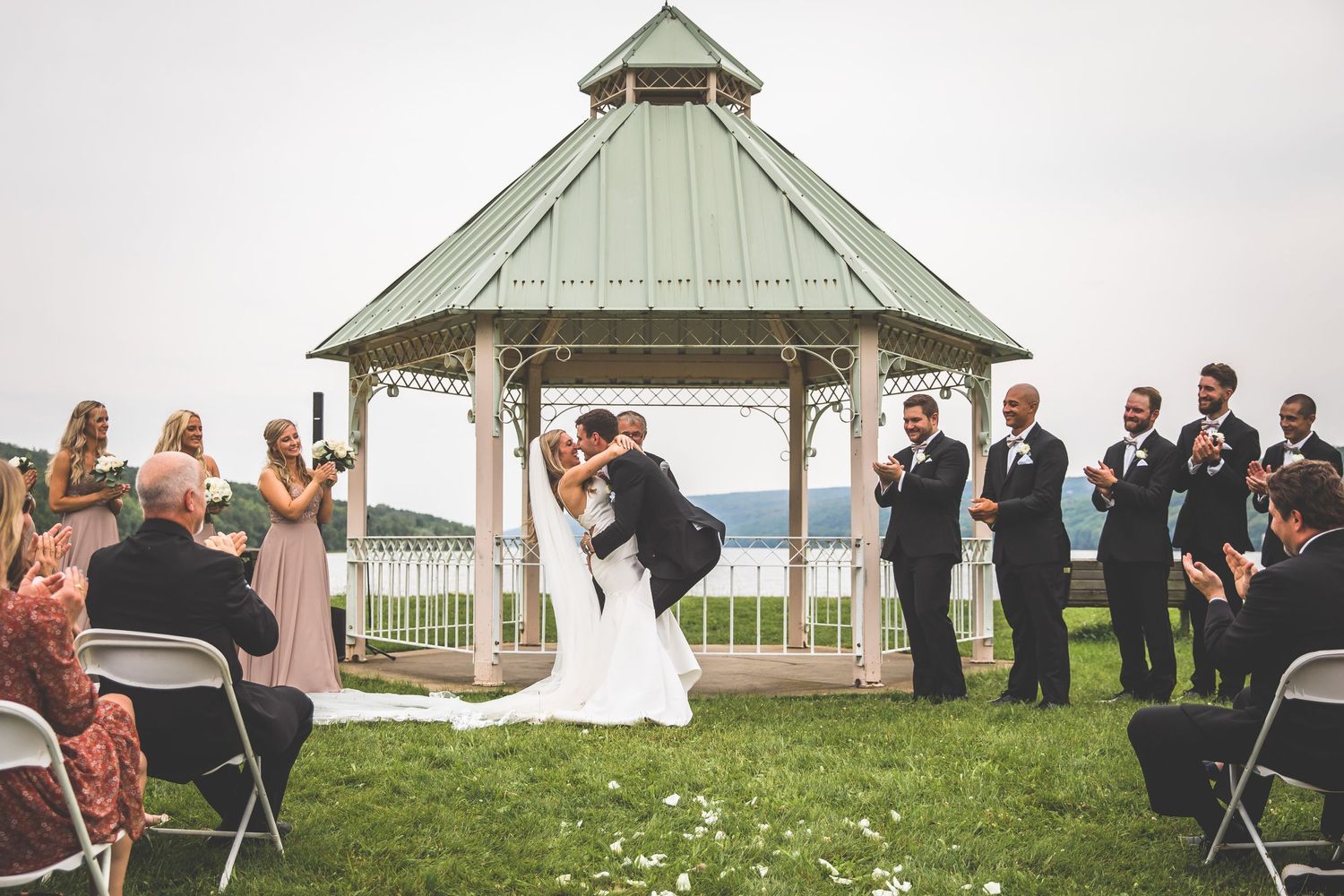 Panoramic shot of wedding guests gathered around a white gazebo for an outdoor lakeside ceremony.