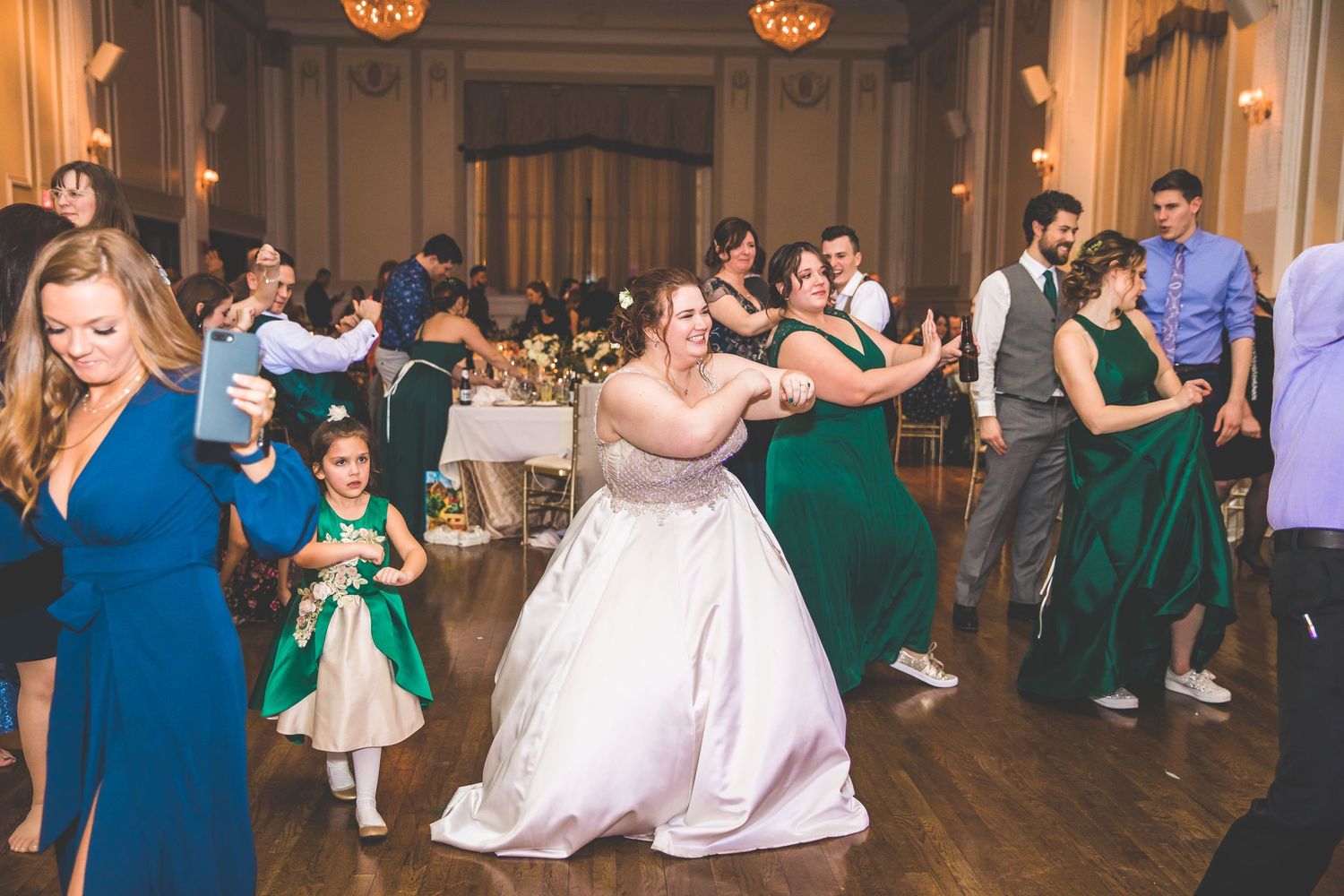 Wedding guests dance beneath warm pendant lights in an elegant ballroom with hardwood floors.