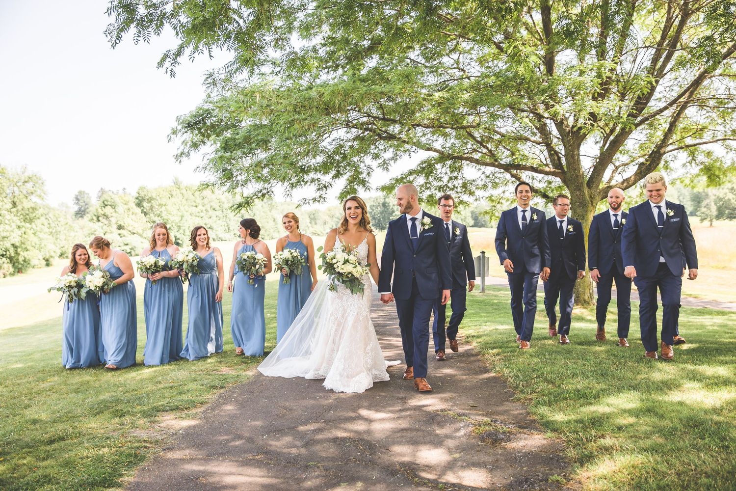 Wedding party poses outdoors in a line with bridesmaids in blue dresses and groomsmen in dark suits.