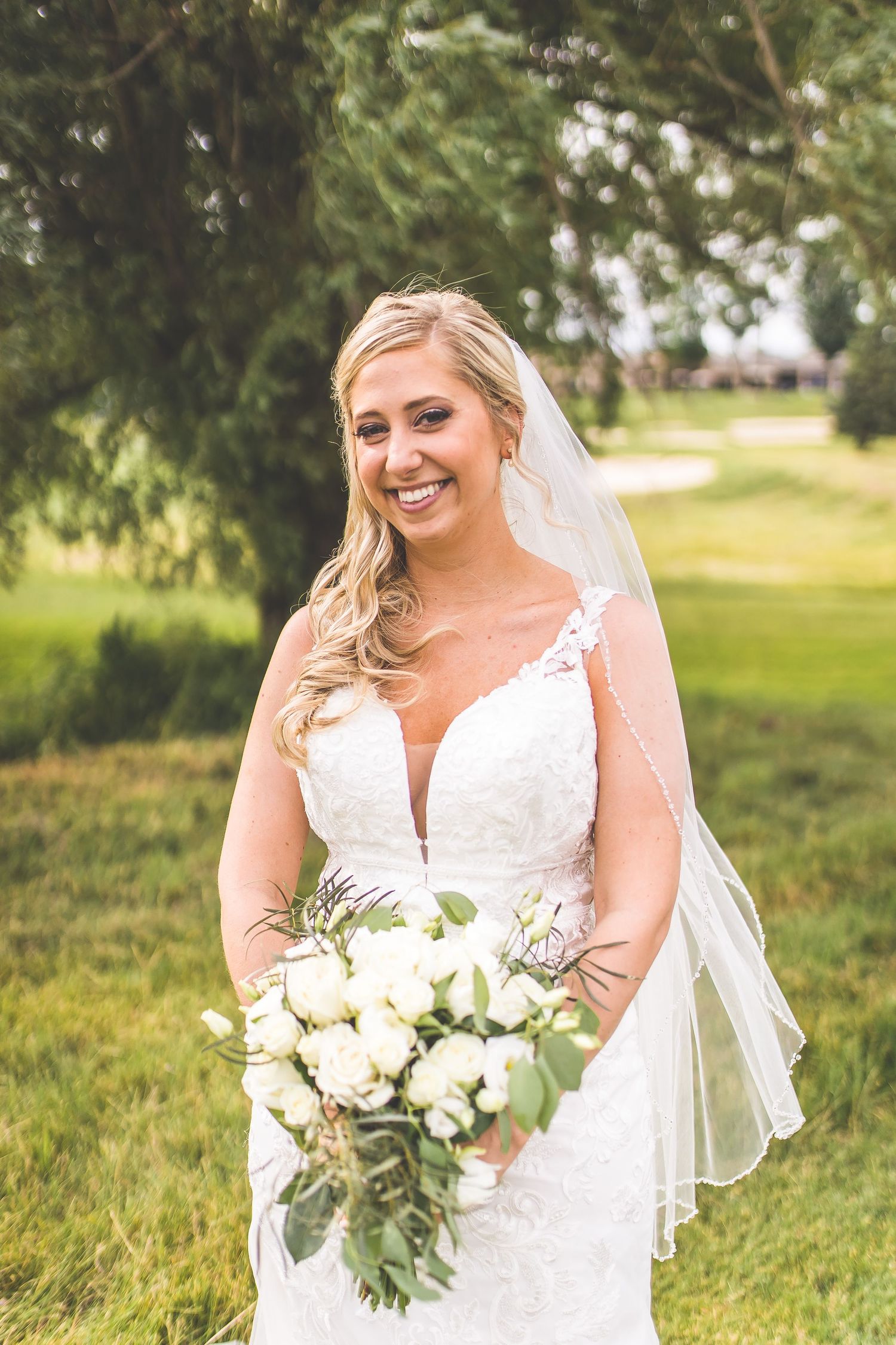 Bride in lace dress holding white bouquet poses in natural outdoor setting.