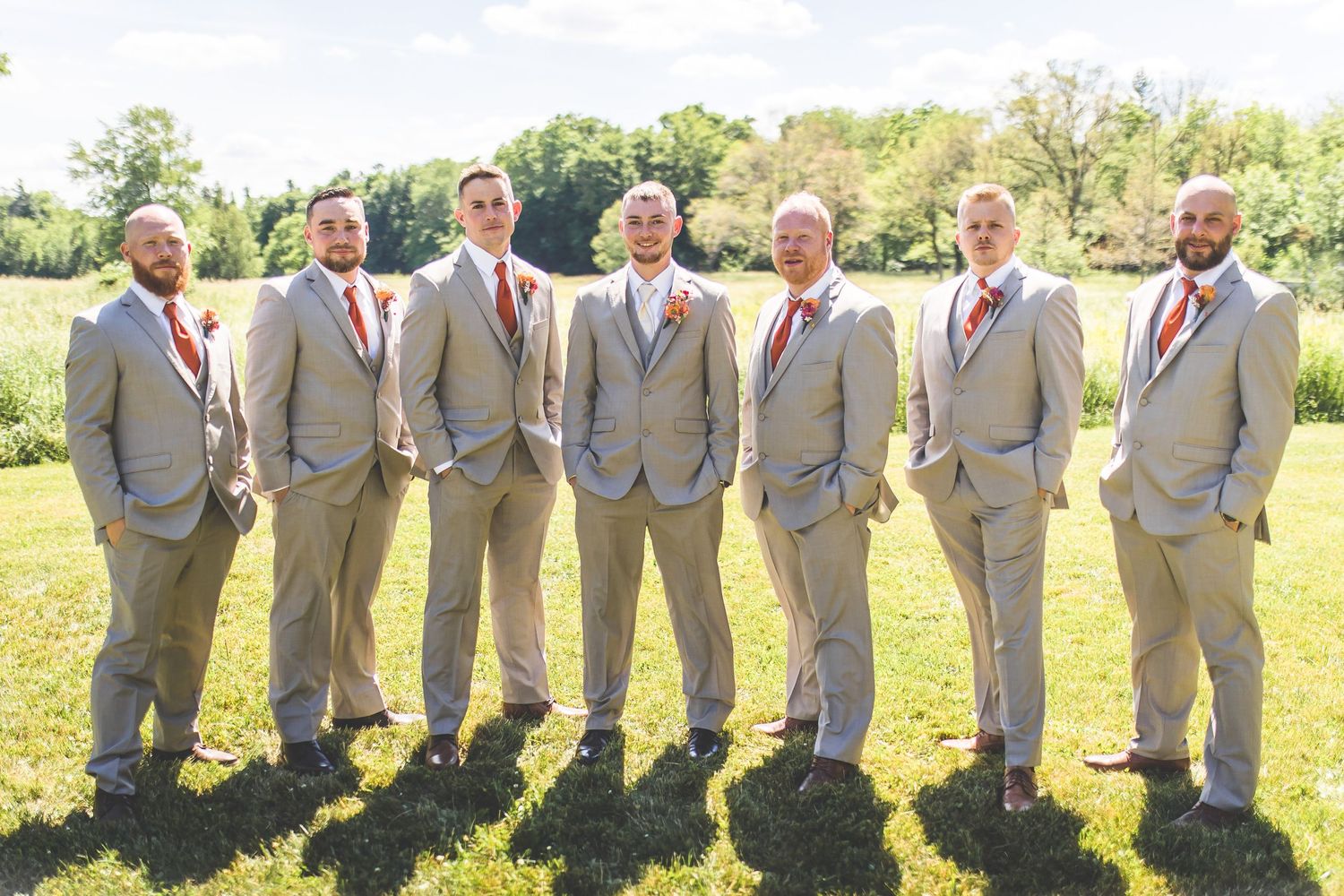 Groomsmen in light gray suits pose together on sunny grass field.