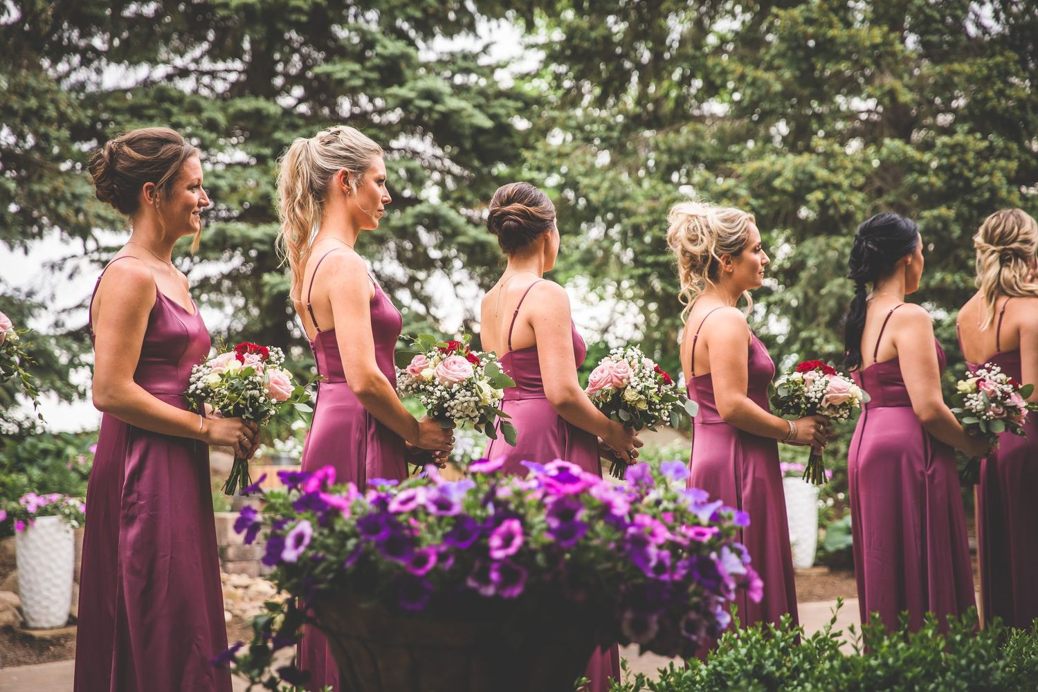 Wedding party wearing burgundy dresses poses outdoors with purple floral bouquets.