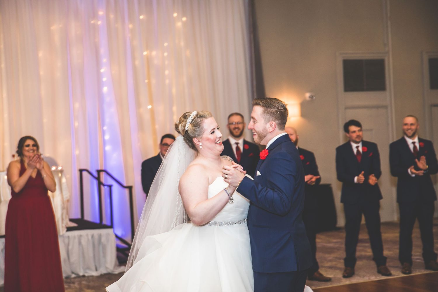 Newlyweds share their first dance at a wedding reception with bridal party watching in the background.