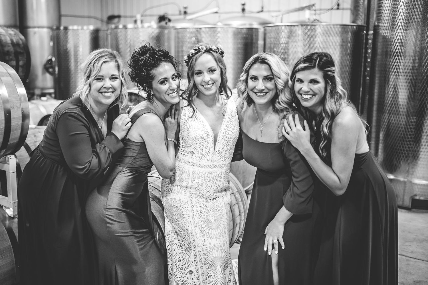 Wedding party celebrates and poses together between rows of wine tanks.