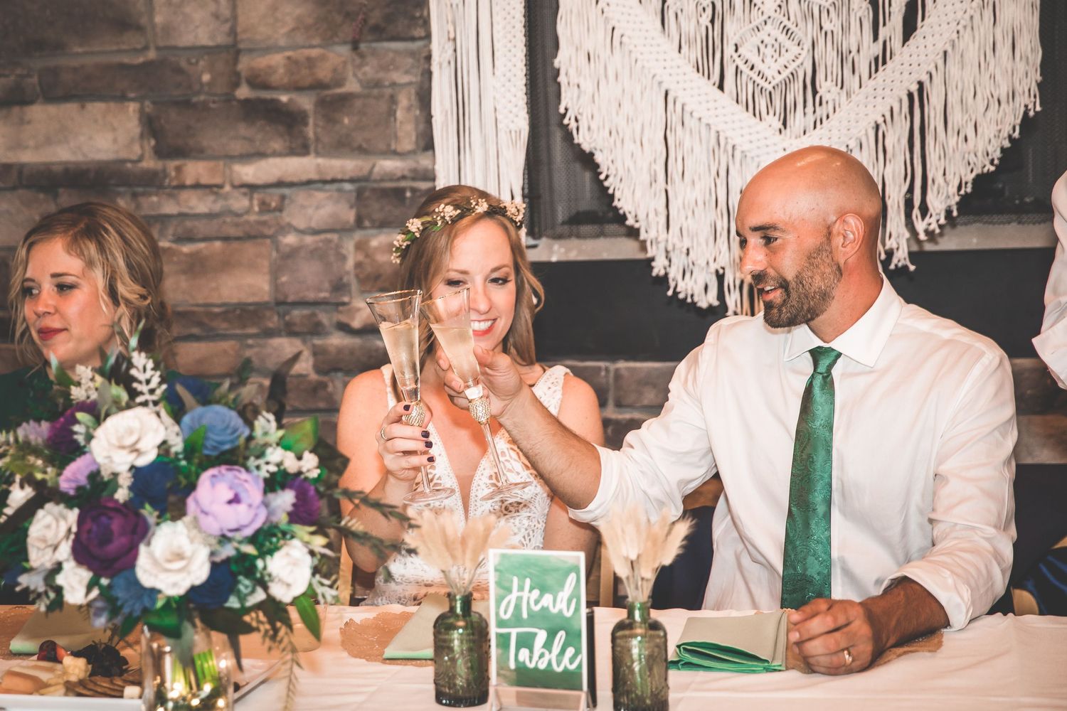 Wedding party seated at head table with elegant decor and stone wall backdrop.