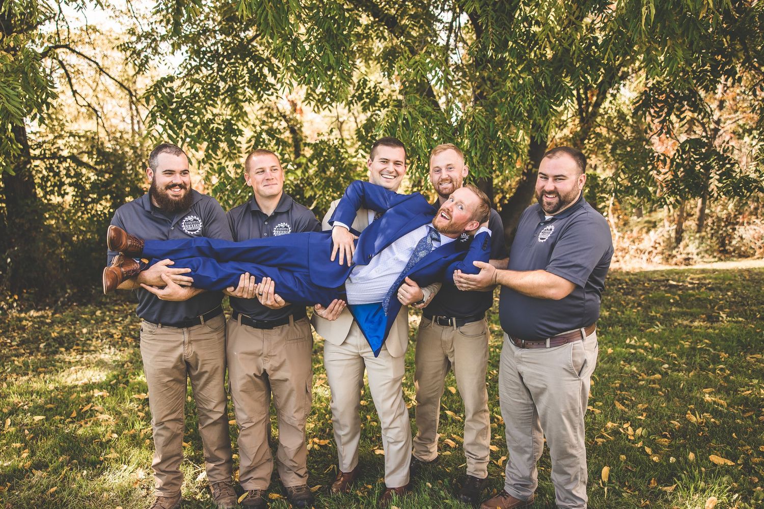 Groomsmen in matching outfits playfully carry groom horizontally for photo.