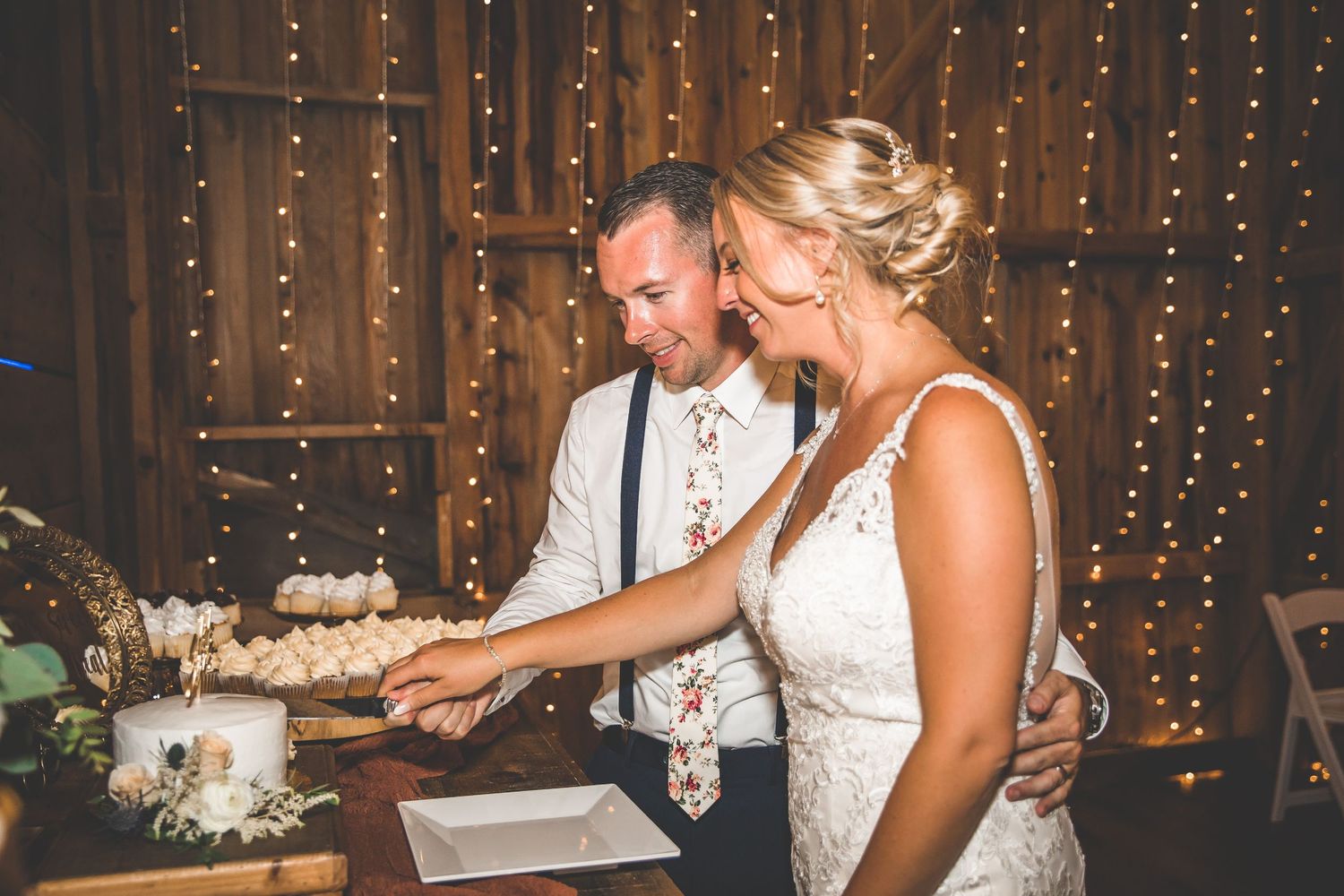 A couple cutting their wedding cake together under string lights in a rustic barn setting.