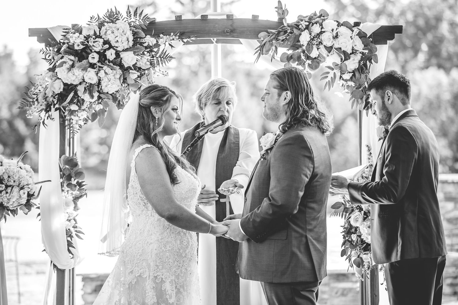 Wedding ceremony taking place outdoors under a white floral arch with guests seated on white chairs.
