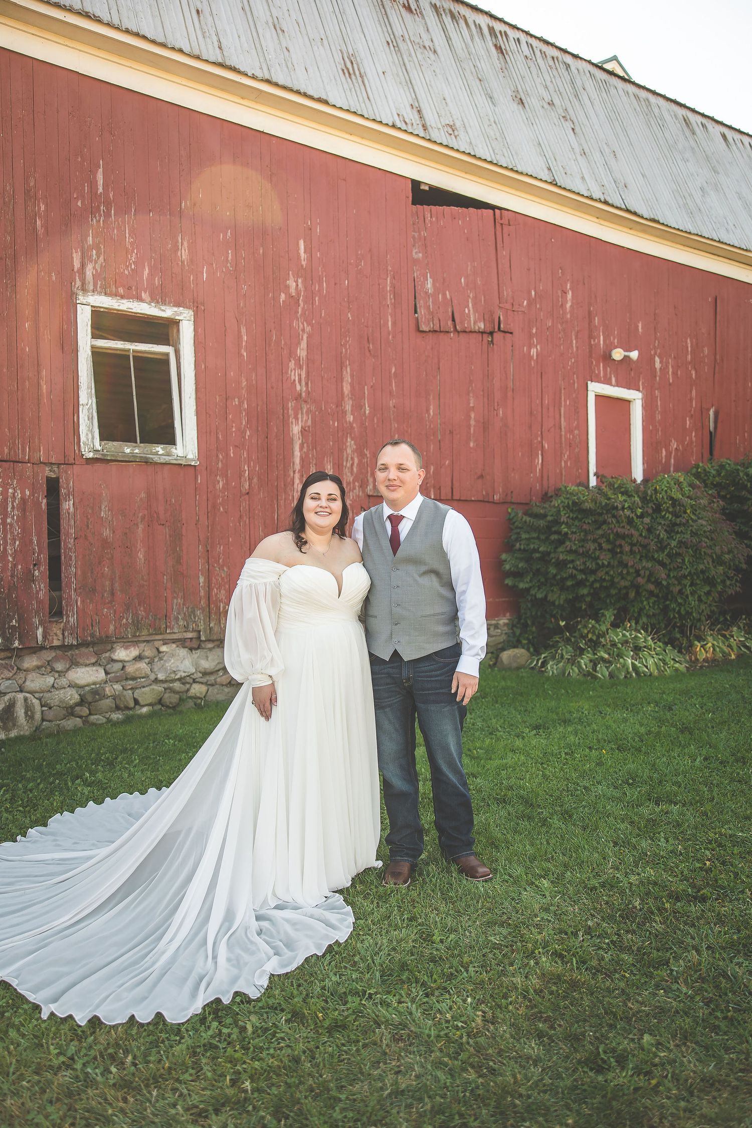 Newlyweds pose together near stone foundation of vintage red barn.