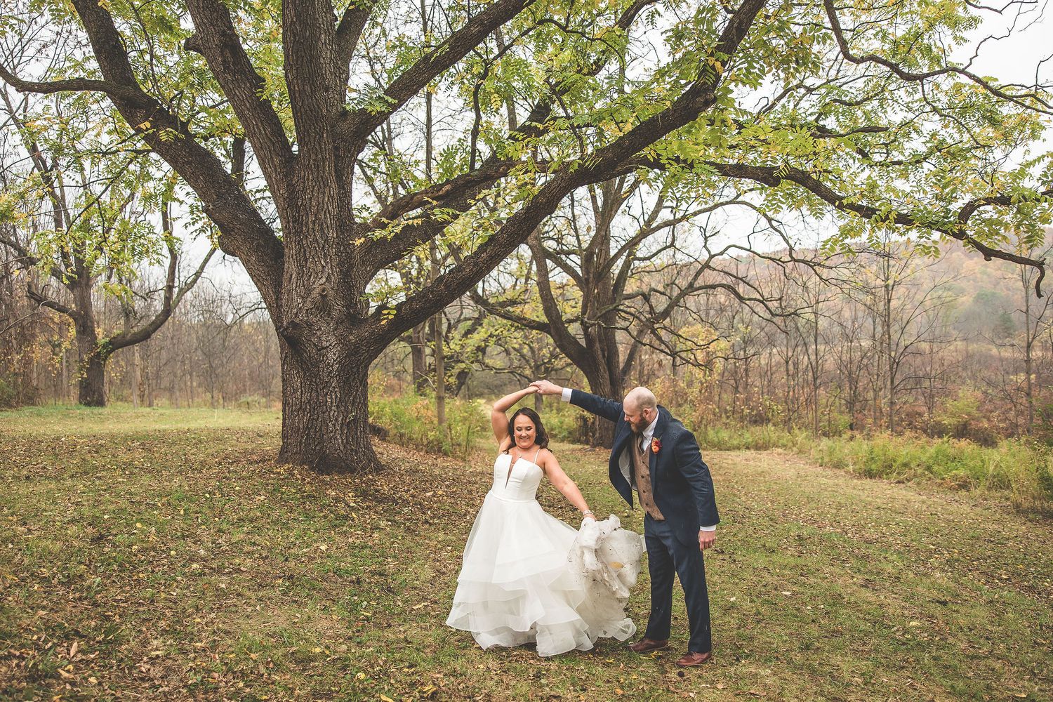 A playful wedding photo with the couple posing under a large tree in a fall landscape.
