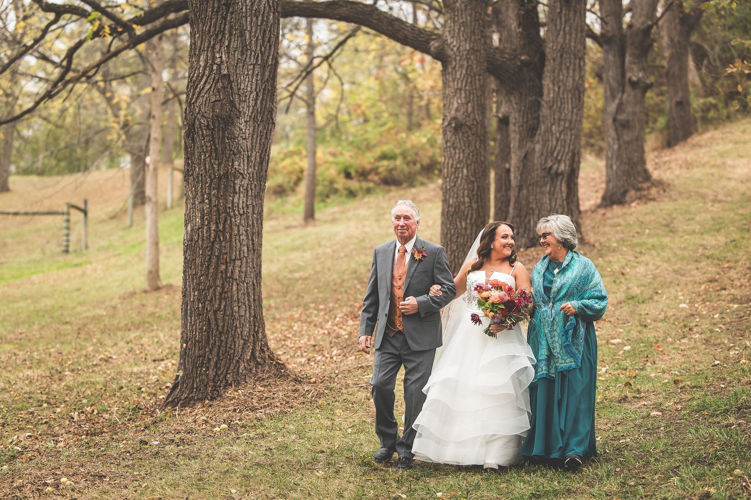 A wedding party walks through a wooded autumn setting with tall trees and fallen leaves.