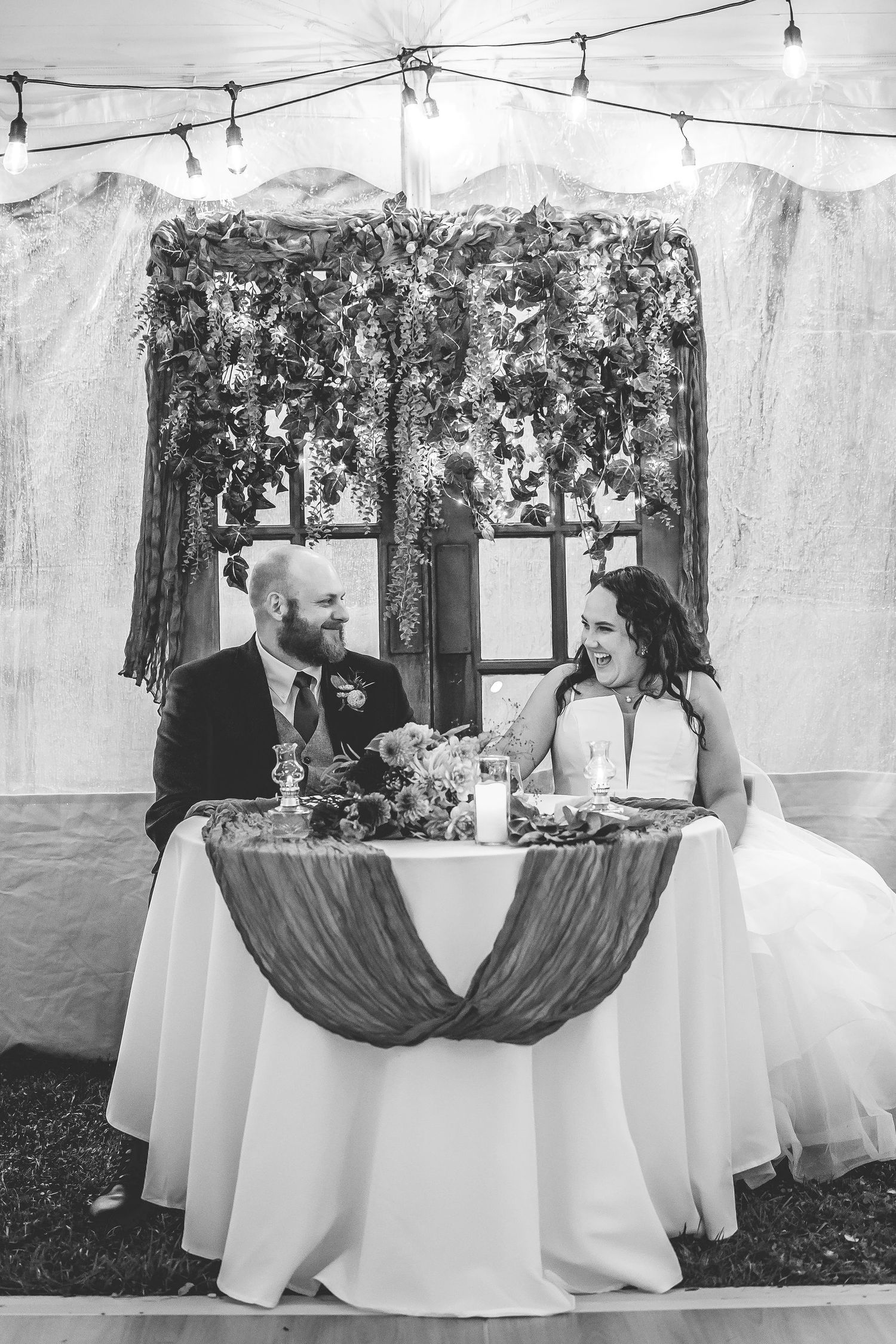 Black and white photo of sweetheart table with burgundy draping and string lights above.
