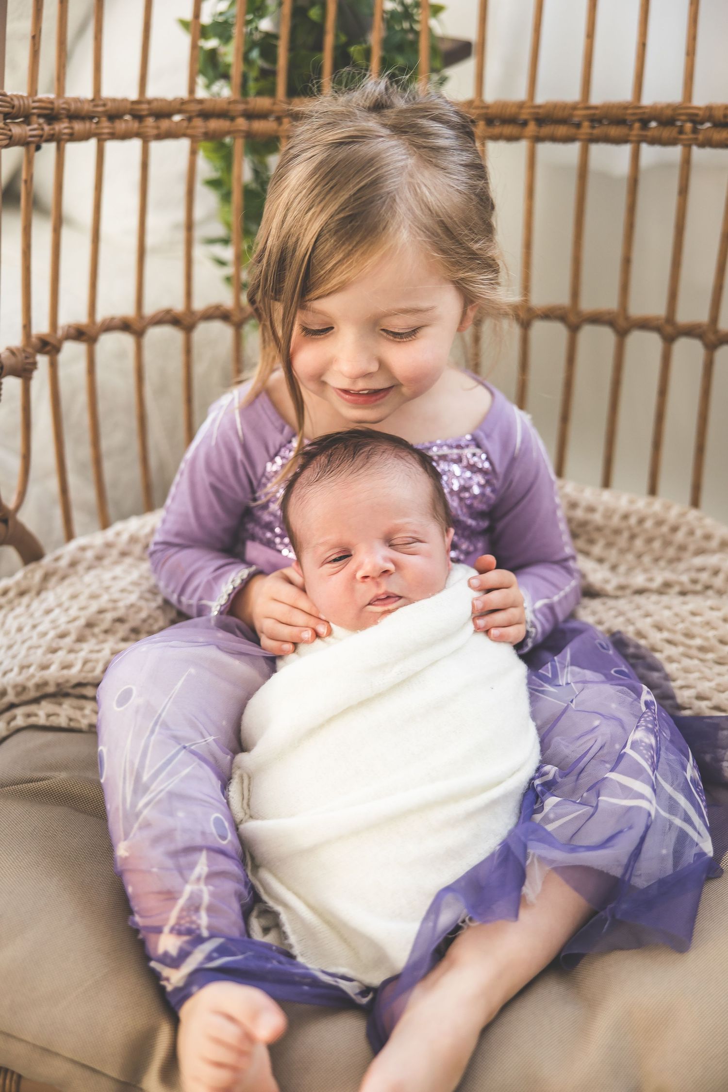 A young child in purple hugs and snuggles with a newborn baby on a couch.