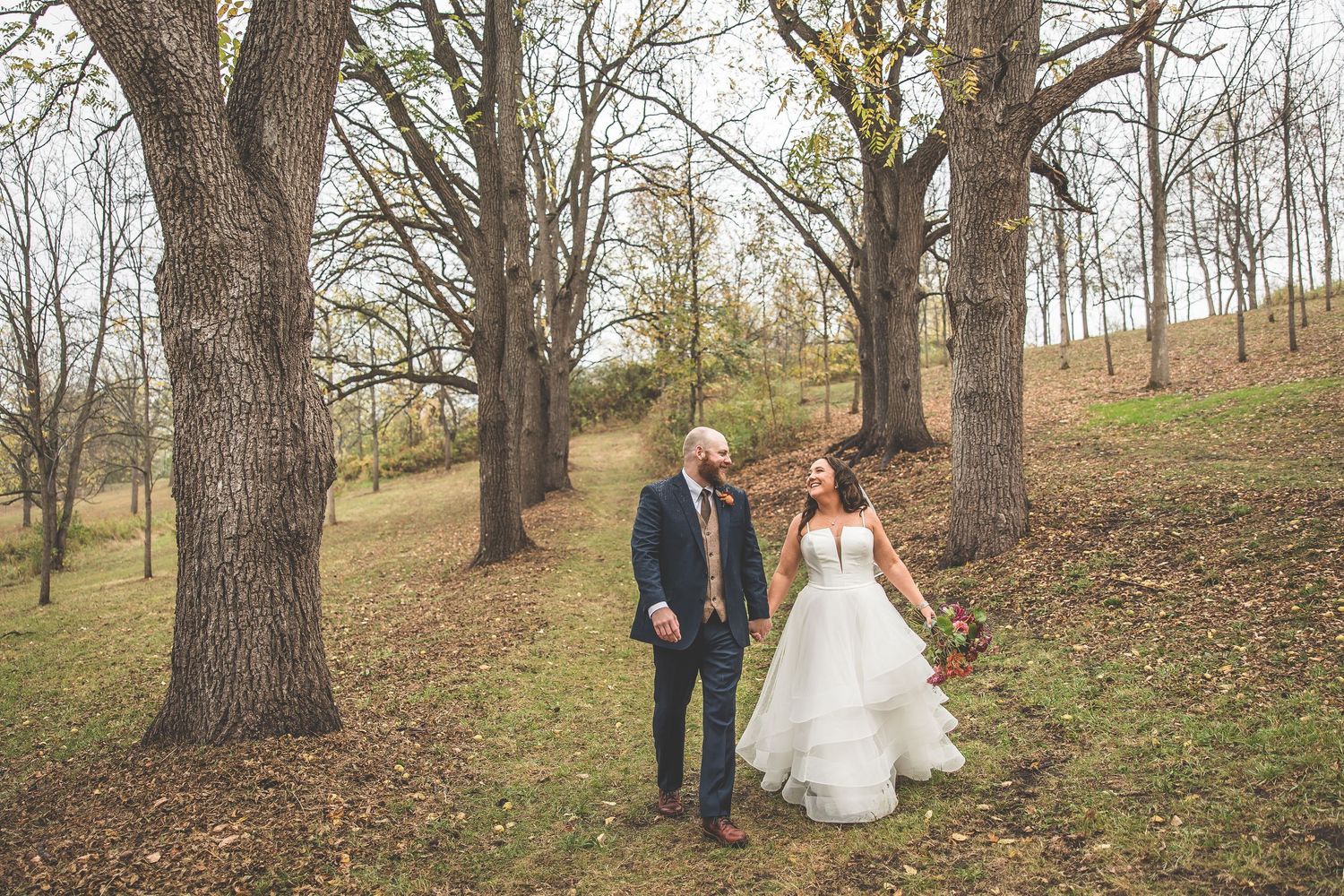 Wedding couple walking through autumn woodland with bare trees and fallen leaves.
