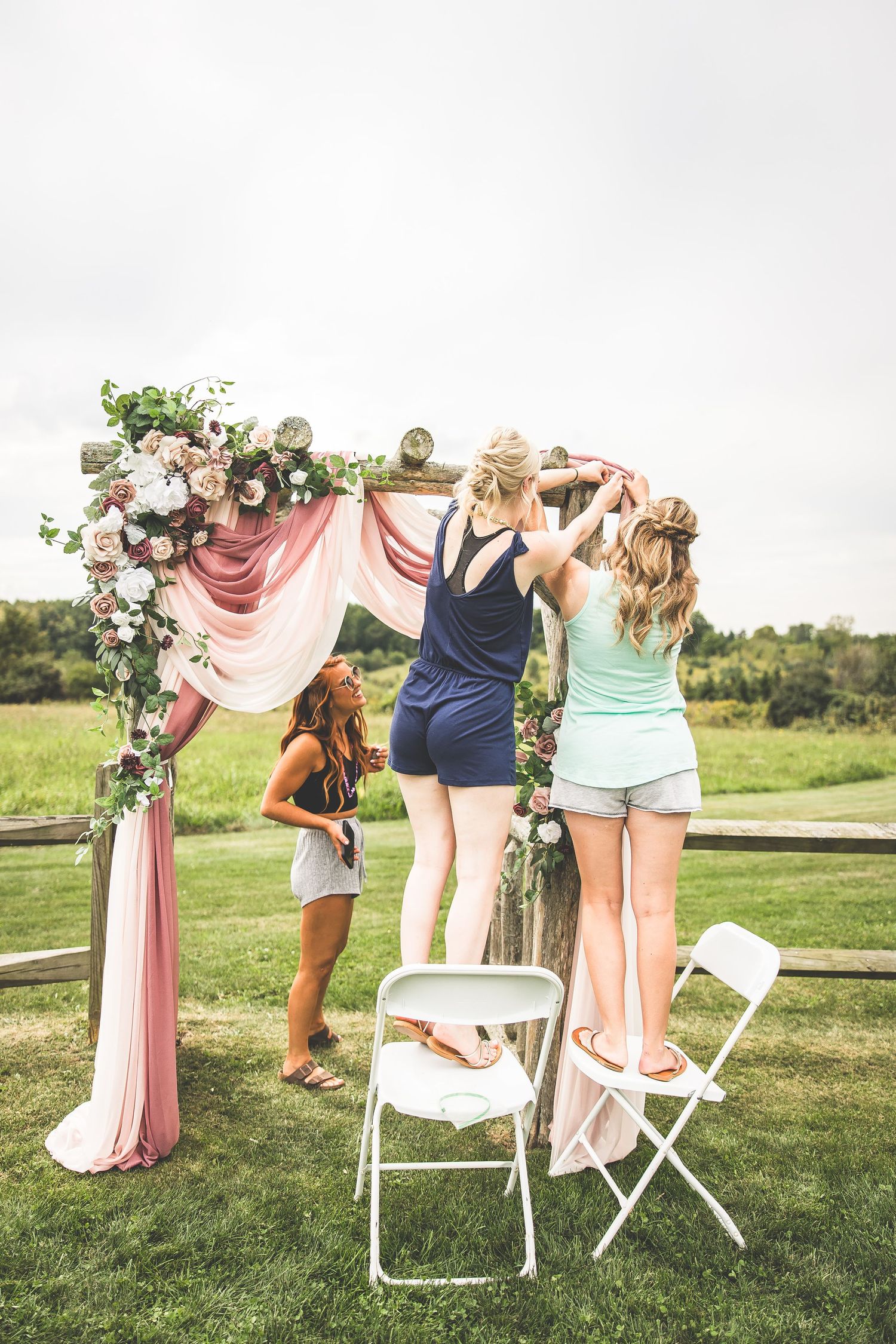 Outdoor wedding setup with white folding chairs and pink fabric decorations in a grassy field.