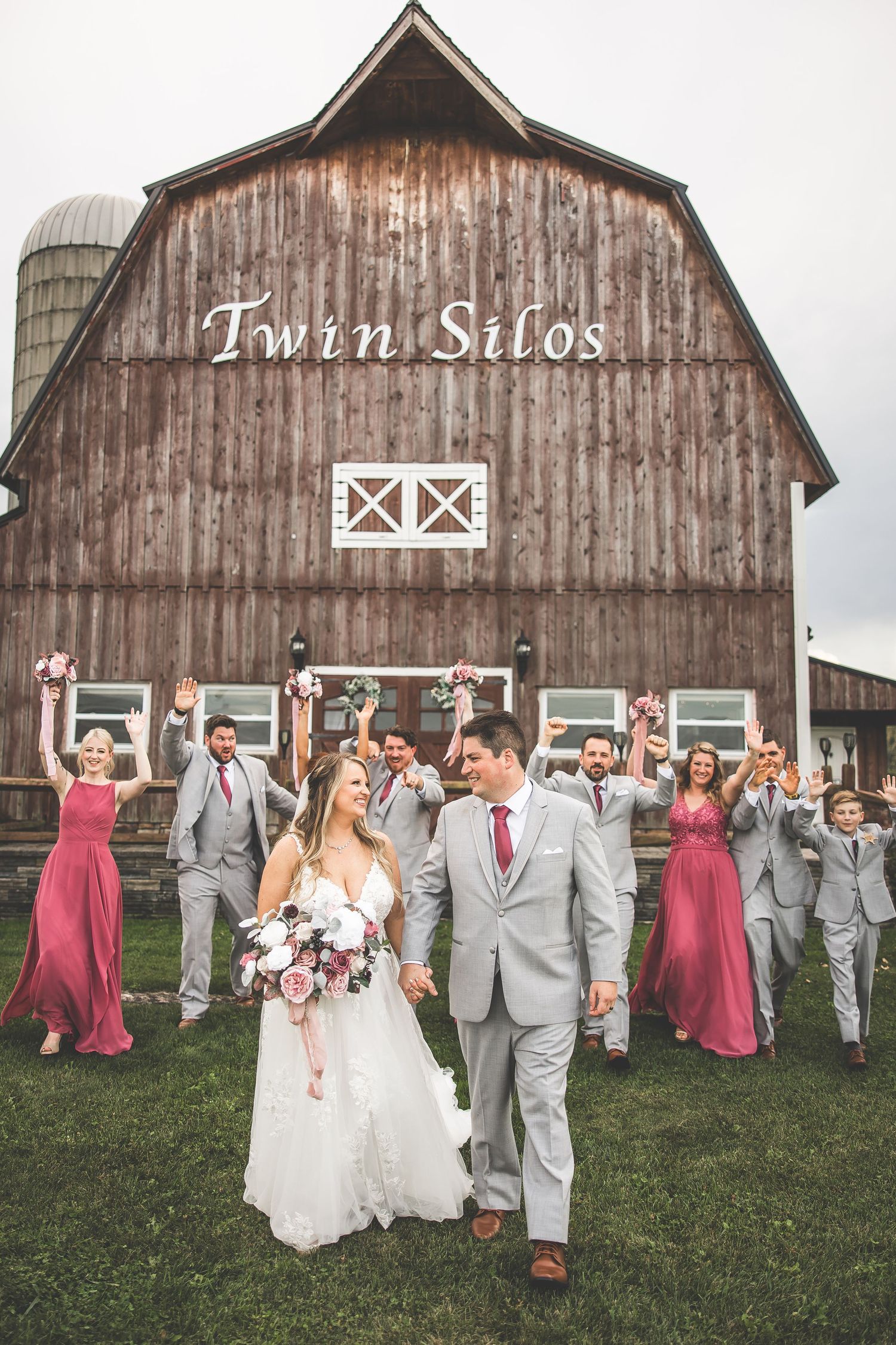 Wedding party poses in front of rustic barn with bridesmaids in burgundy dresses and groomsmen in gray suits.