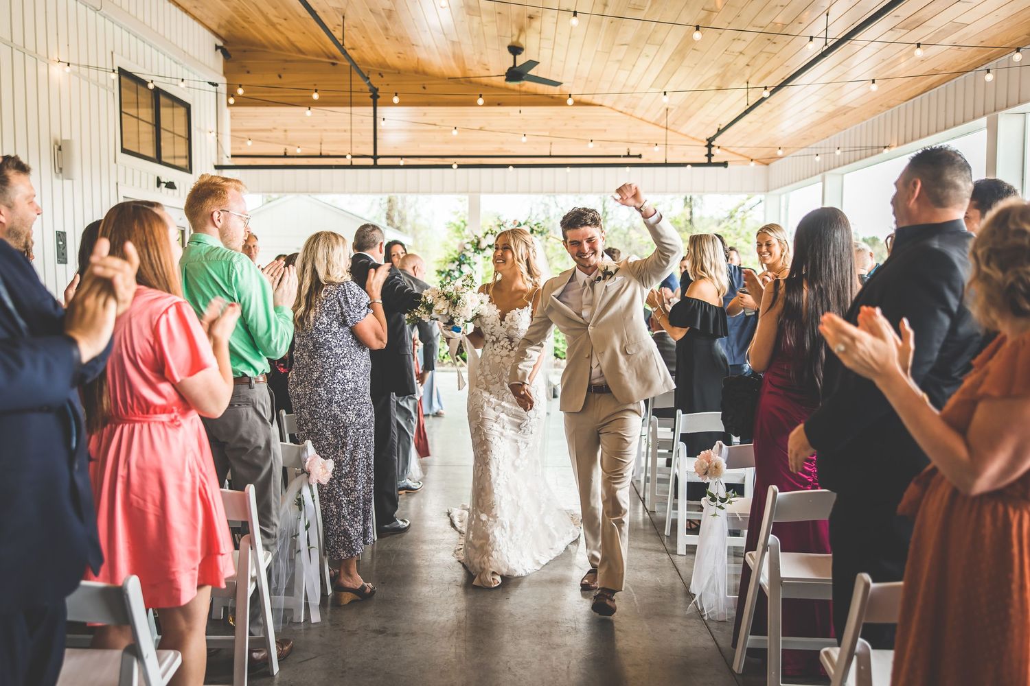 Wedding guests stand and applaud during ceremony in rustic indoor venue.