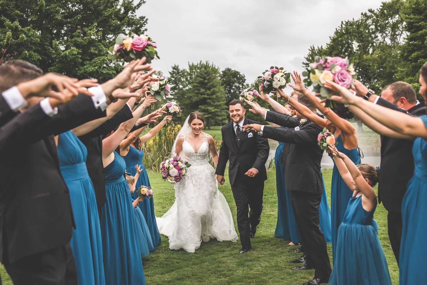 Wedding party creates tunnel formation for newlyweds to walk through on grassy lawn.