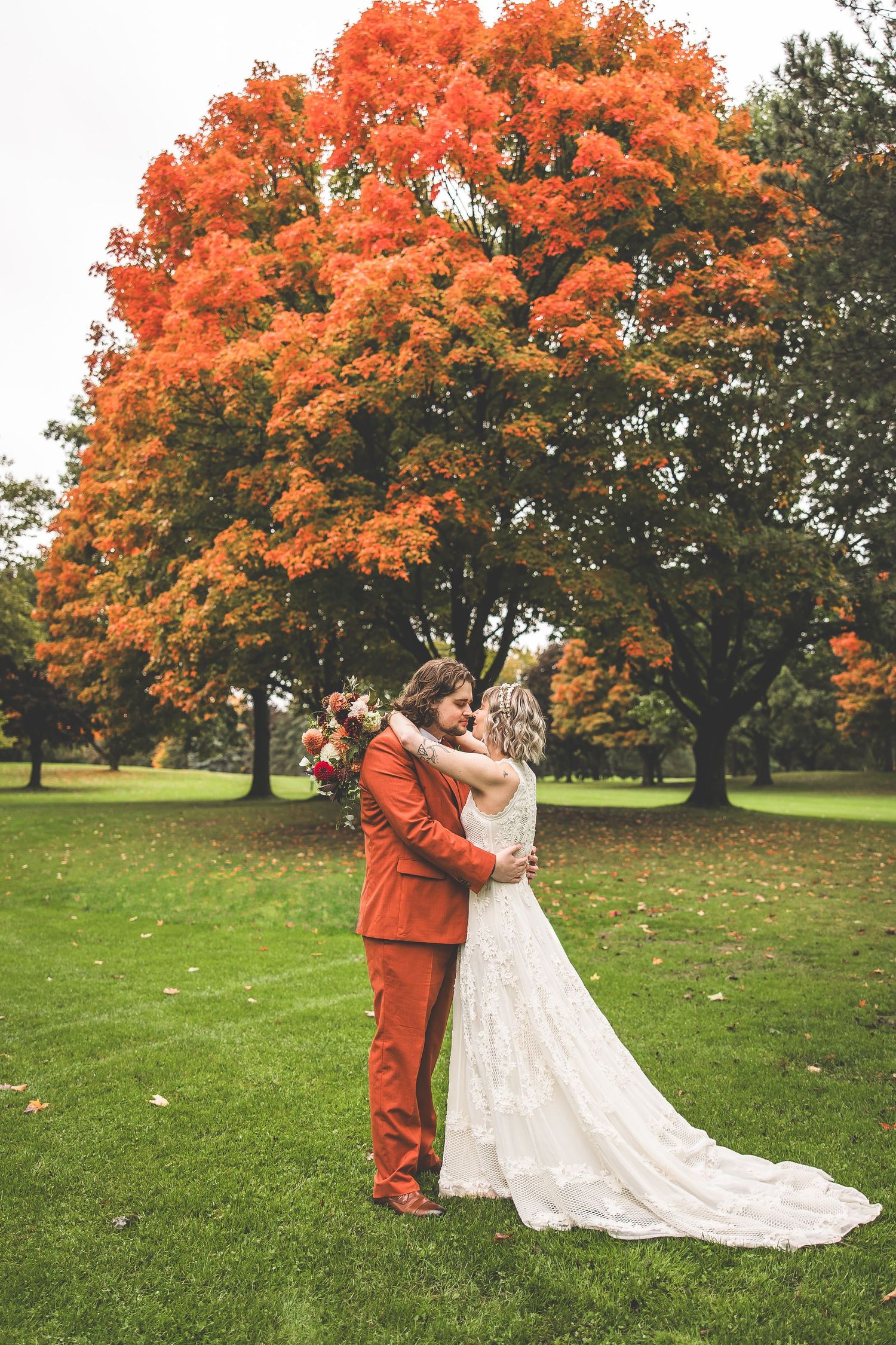 A couple embraces on a lawn beneath a stunning orange maple tree during their fall wedding.